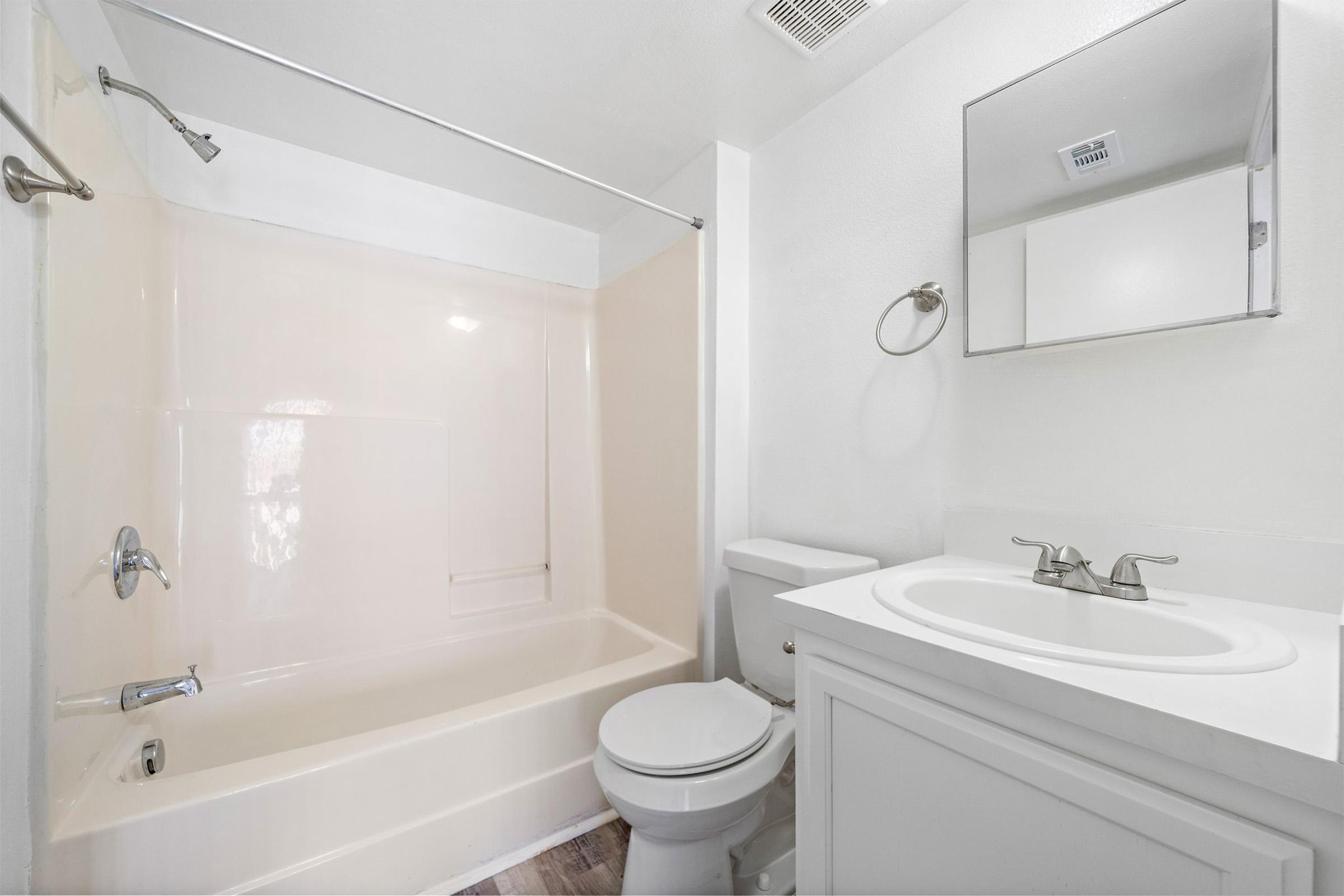 A clean and neutral-toned bathroom featuring a bathtub with a shower, a toilet, and a vanity with a sink. The walls are painted white, and there is a simple mirror above the sink. The flooring appears to be a light wood design, creating a bright and inviting space.