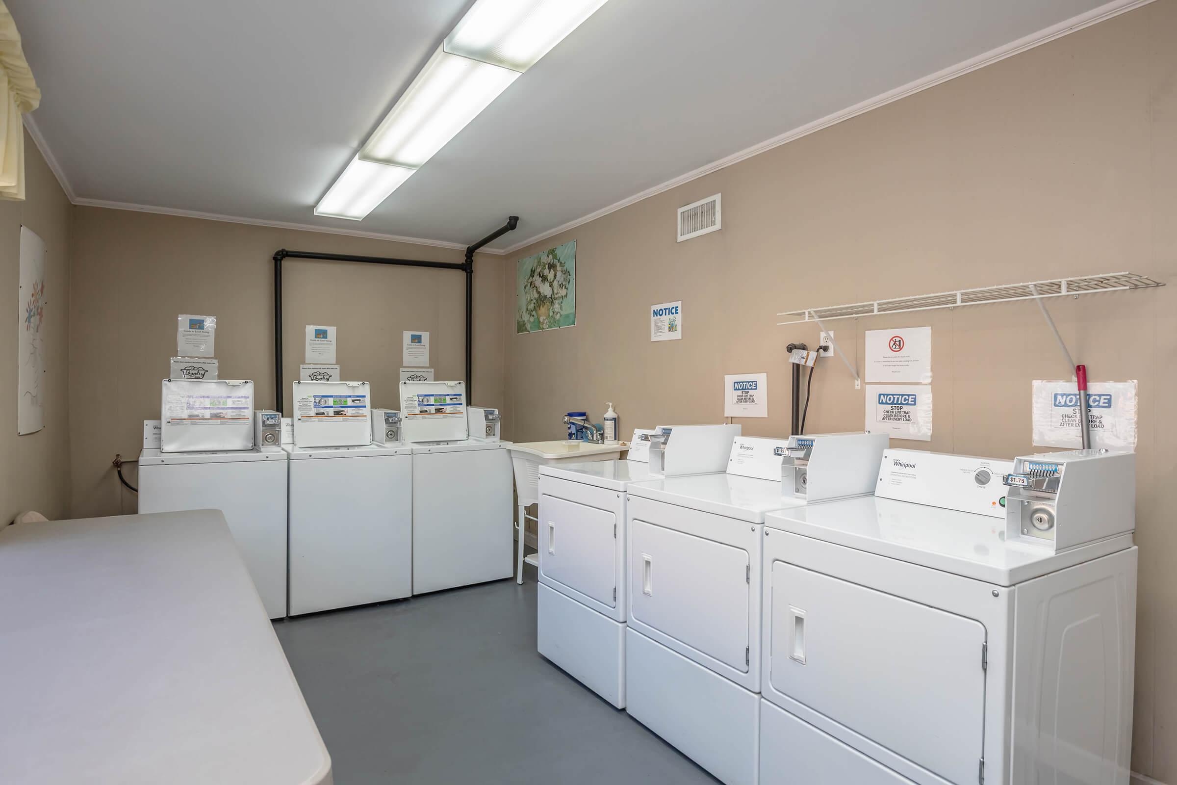 A clean, well-lit laundry room featuring several white washing machines and dryers lined against the walls. There are shelves for supplies, notice signs on the machines, and a small table for folding laundry. The walls are painted a neutral color, creating a tidy and organized space.