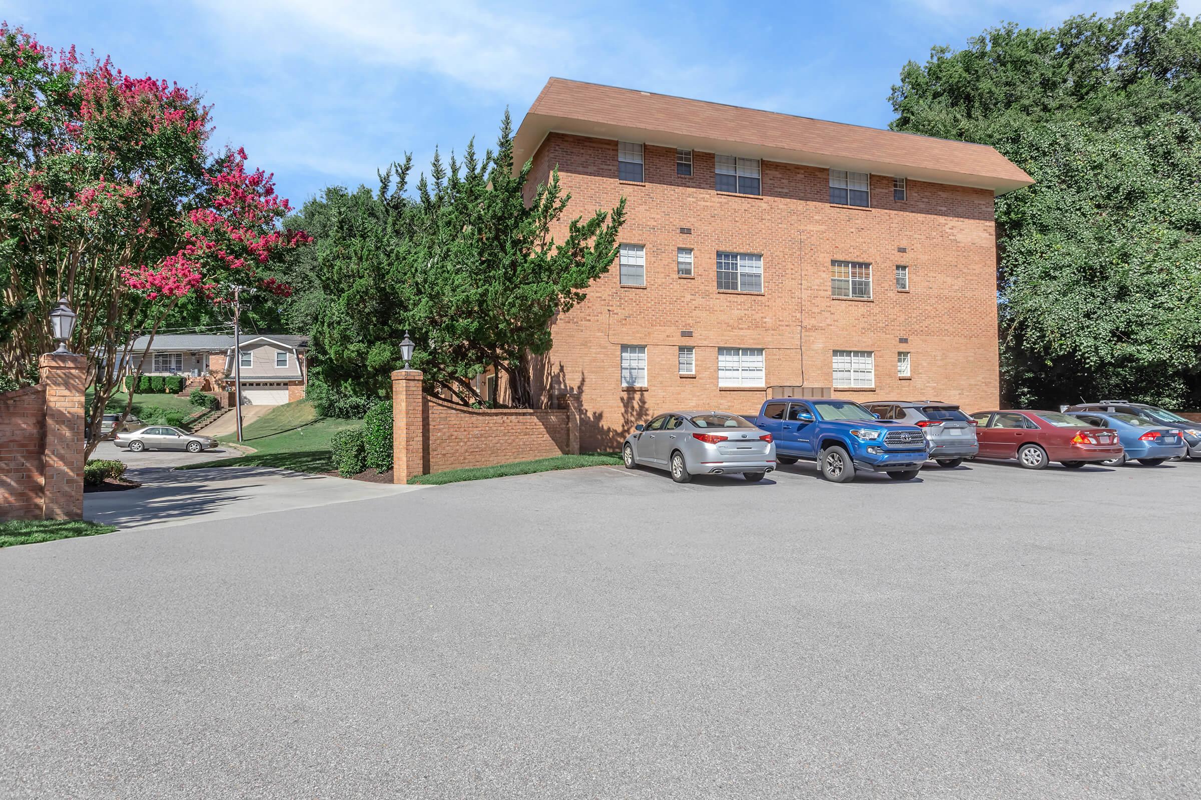 A brick apartment building surrounded by trees and shrubs, with a parking lot in front featuring several parked cars. The sky is clear and blue, indicating a sunny day. A smaller building is visible in the background, enhancing the residential setting.