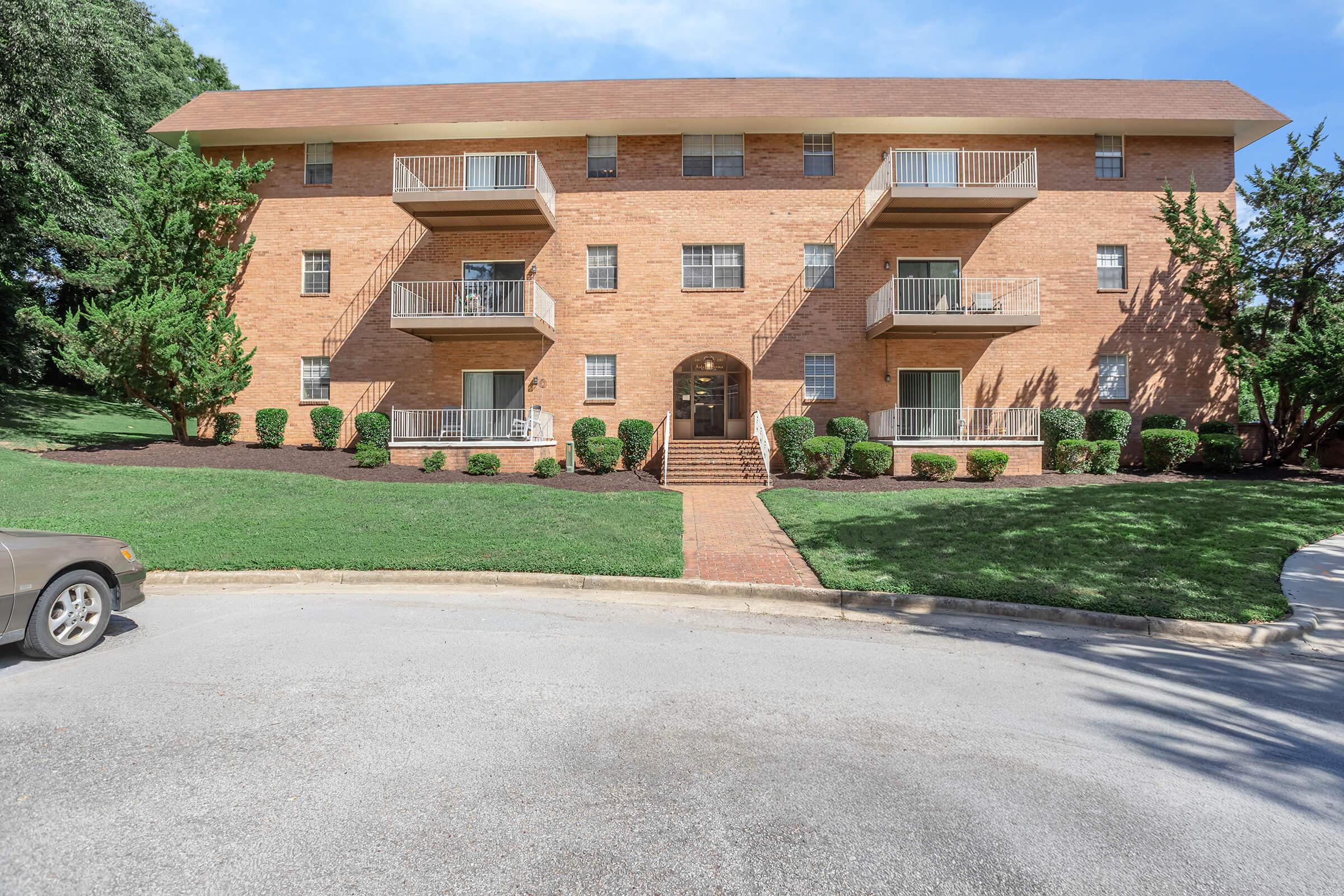 Brick apartment building with four levels, featuring balconies on each floor. The building is surrounded by neatly trimmed bushes and grass, with a walkway leading to the main entrance. A circular driveway is visible in front of the building, along with a parked car in the foreground.