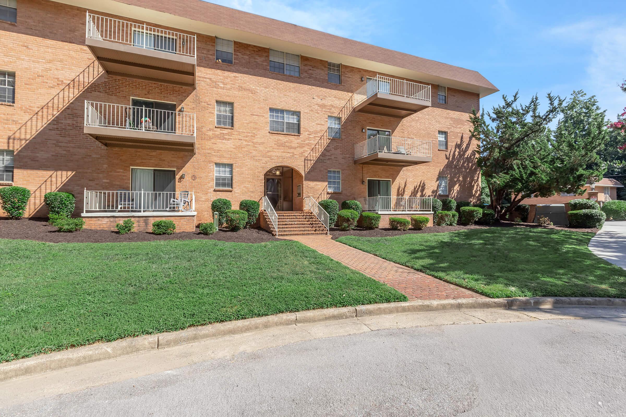 A brick apartment building with multiple balconies and landscaped lawns in front. The building features a clear blue sky and well-maintained greenery, including bushes and a small tree. A curved driveway leads to the entrance, enhancing the inviting appearance of the property.