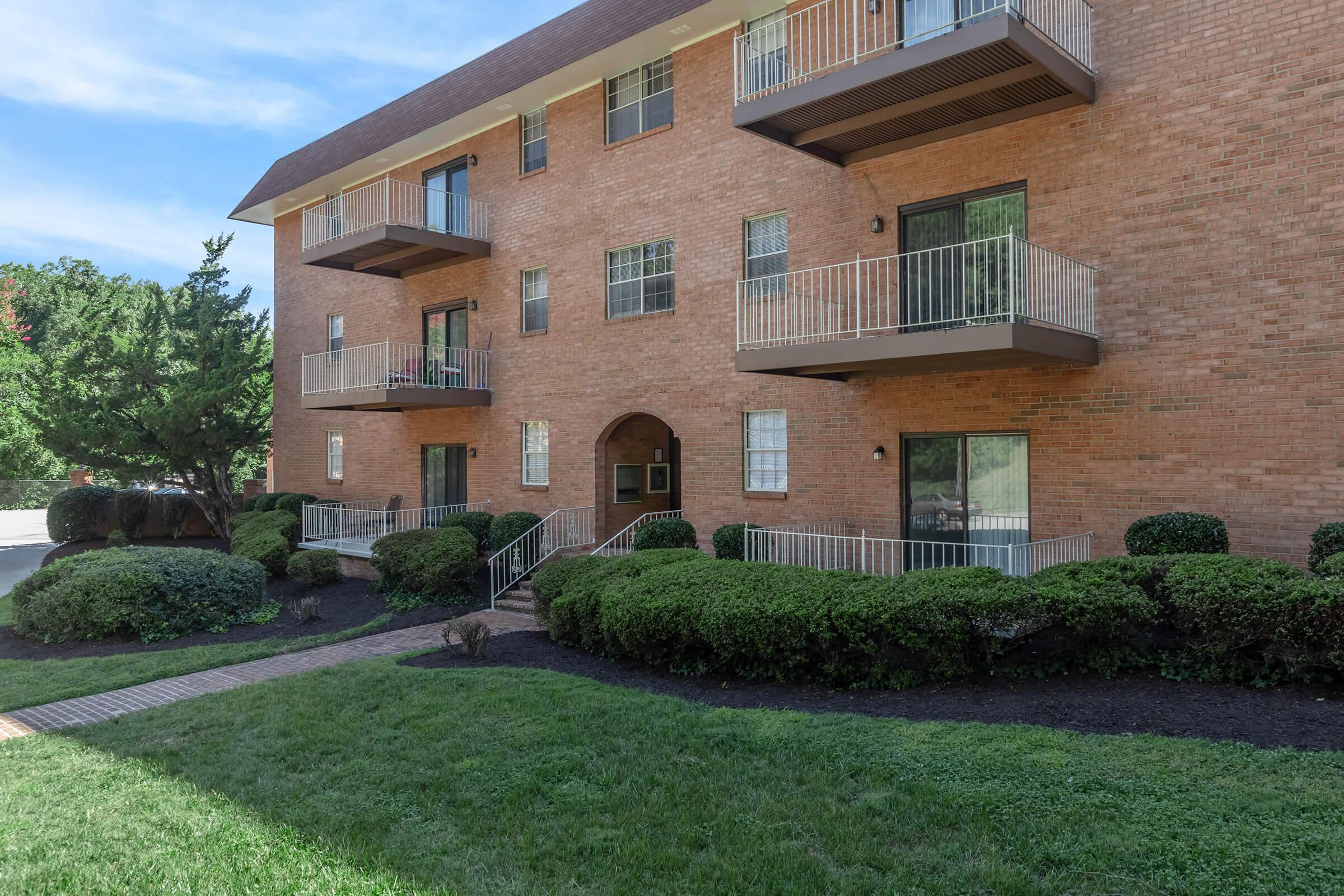 A brick apartment building with multiple balconies, surrounded by well-manicured shrubbery and a small lawn. The scene features a clear blue sky and a pathway leading to the entrance of the building.