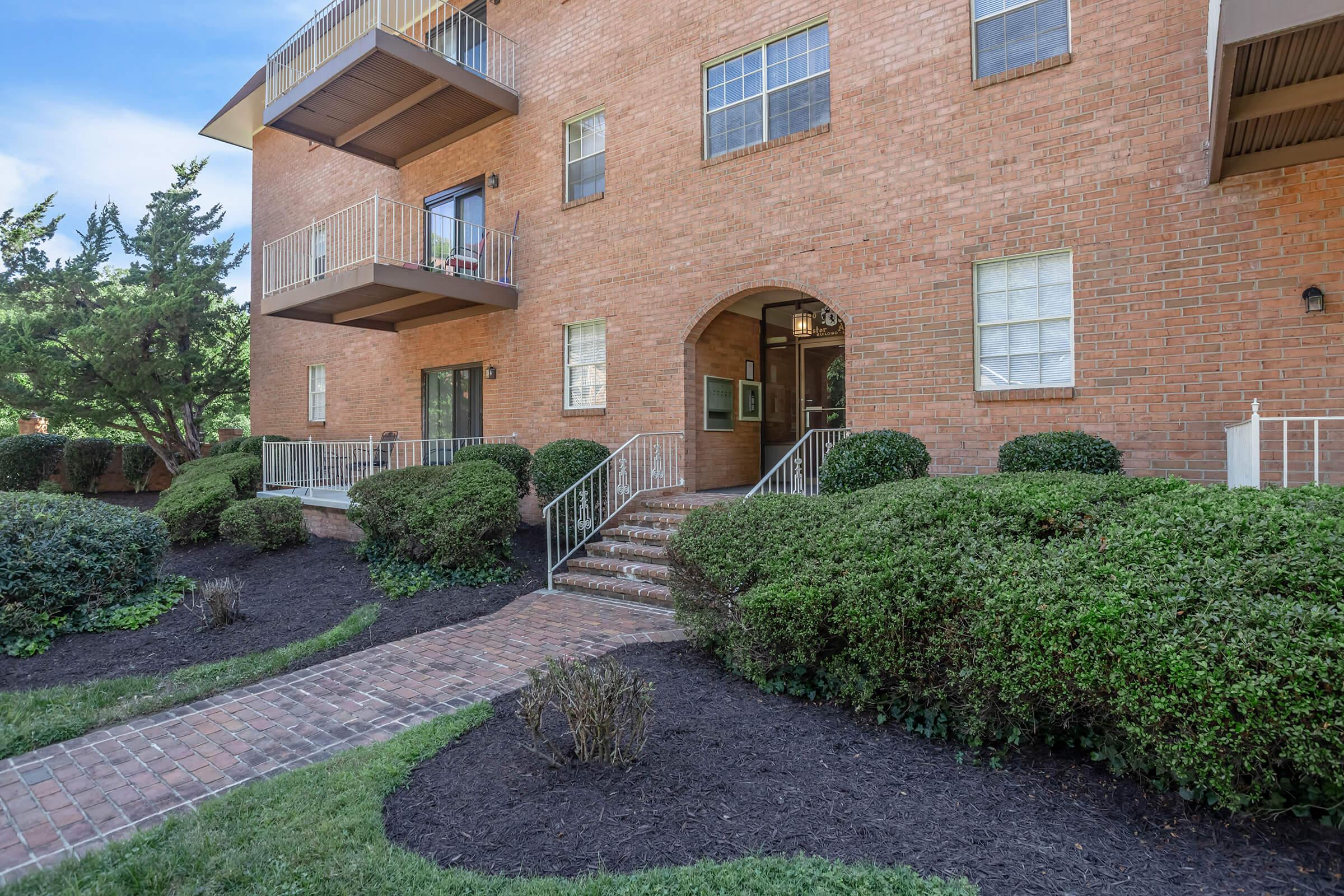 A brick apartment building featuring a set of stairs leading to an arched entrance. Surrounding the entry are neatly trimmed hedges and a brick pathway, with additional greenery visible in the area. The building has multiple balconies and large windows.
