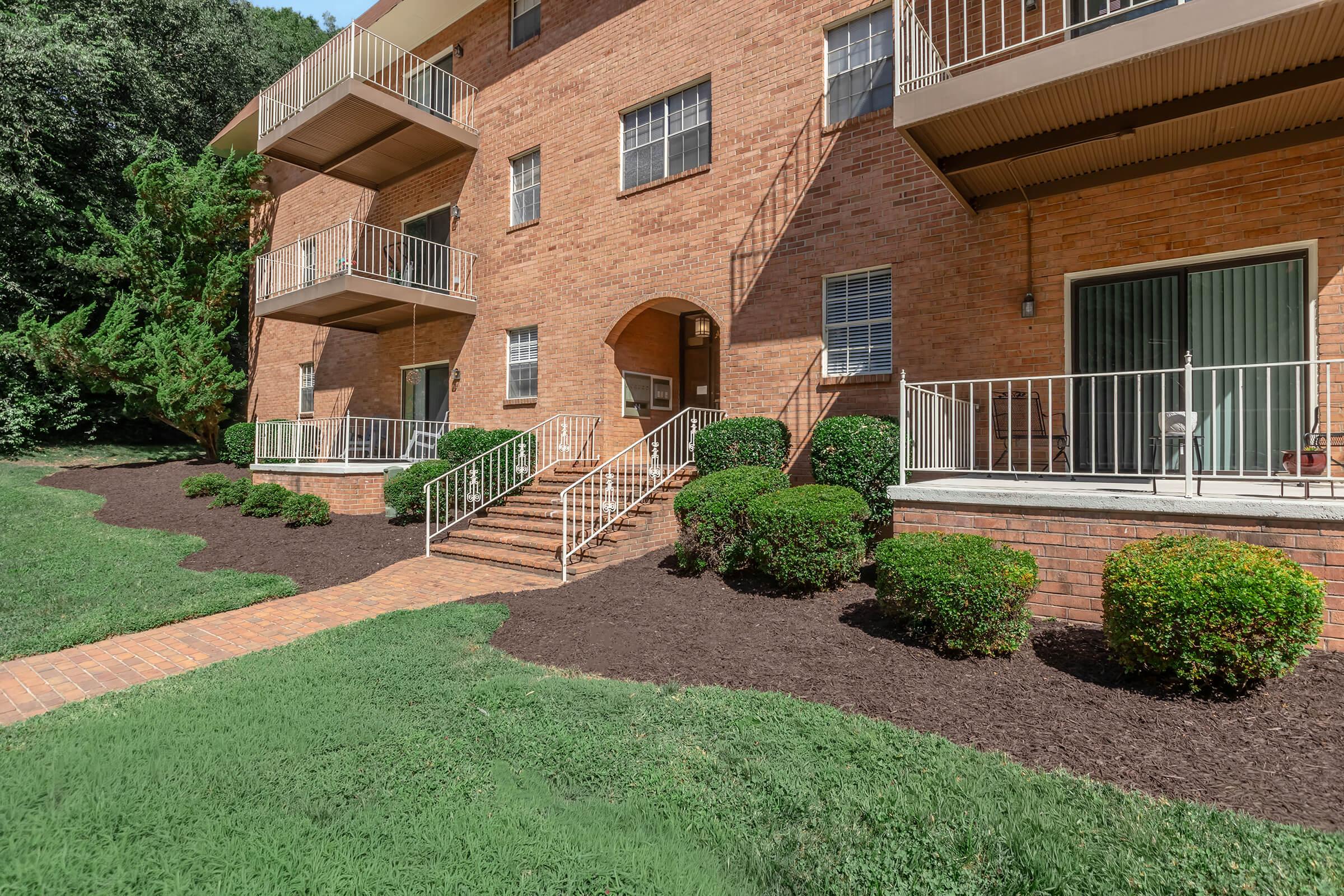 View of a brick apartment building with three levels, featuring balconies and an entrance with stairs leading up. The landscaping includes well-trimmed bushes and a paved walkway, set against a backdrop of green grass and trees. Sunlight casts shadows on the building's façade.