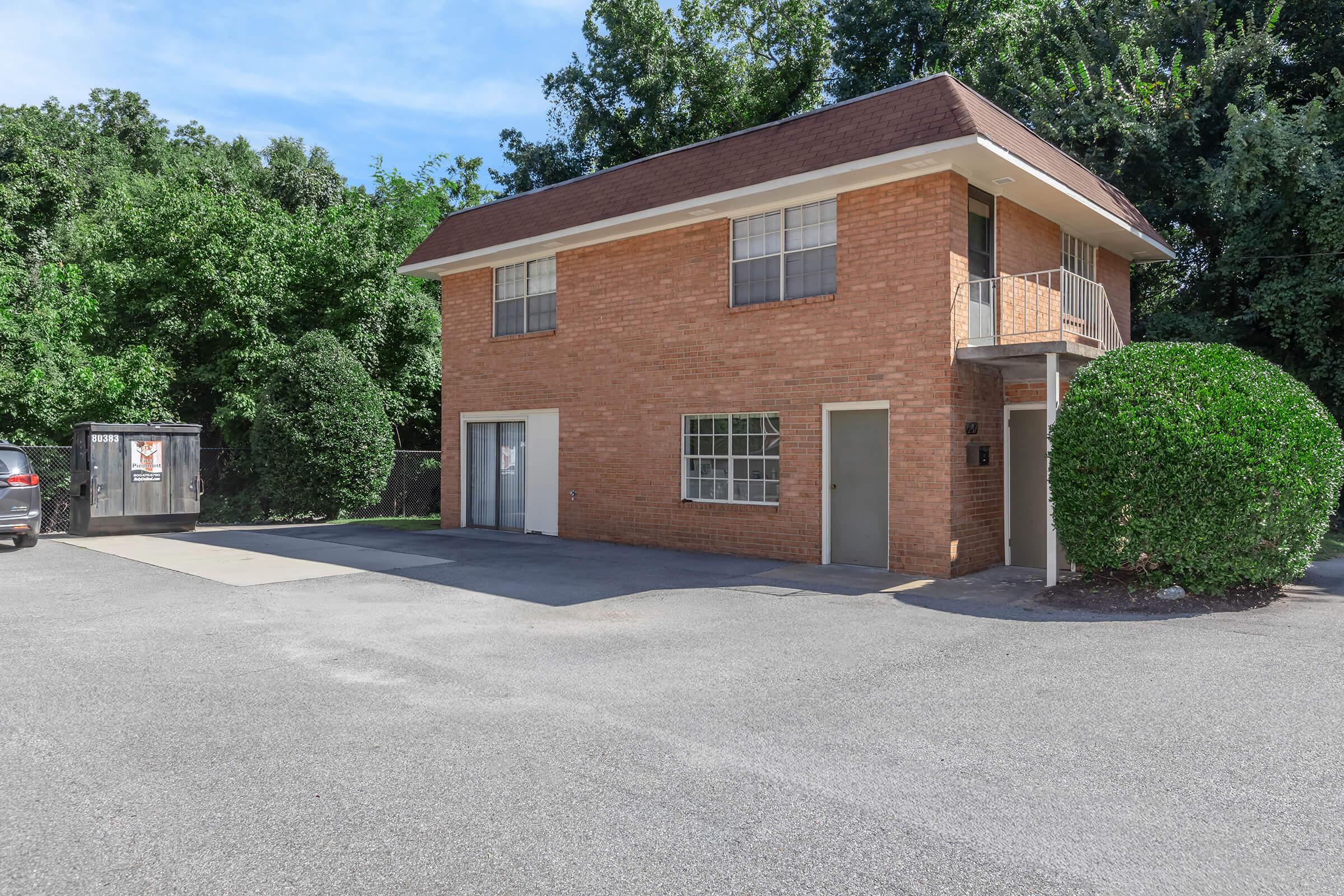 A two-story brick building with a flat roof, featuring several windows and a staircase on the right side. It is surrounded by greenery and has a paved parking area with a dumpster nearby. The scene is set on a sunny day with clear blue skies.