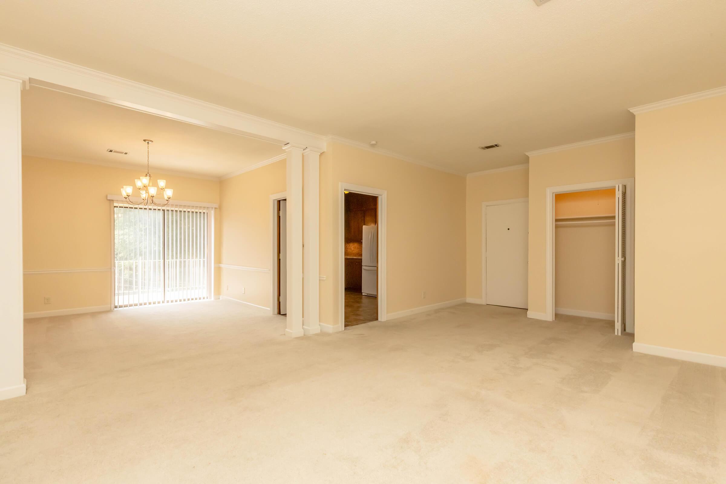 Spacious, empty living room featuring beige walls and carpet, with a chandelier visible in the background. Sunlight streams through vertical blinds, illuminating the area. A doorway leads to another room on the right, while a closet is partially visible in the corner. The overall ambiance is bright and airy.