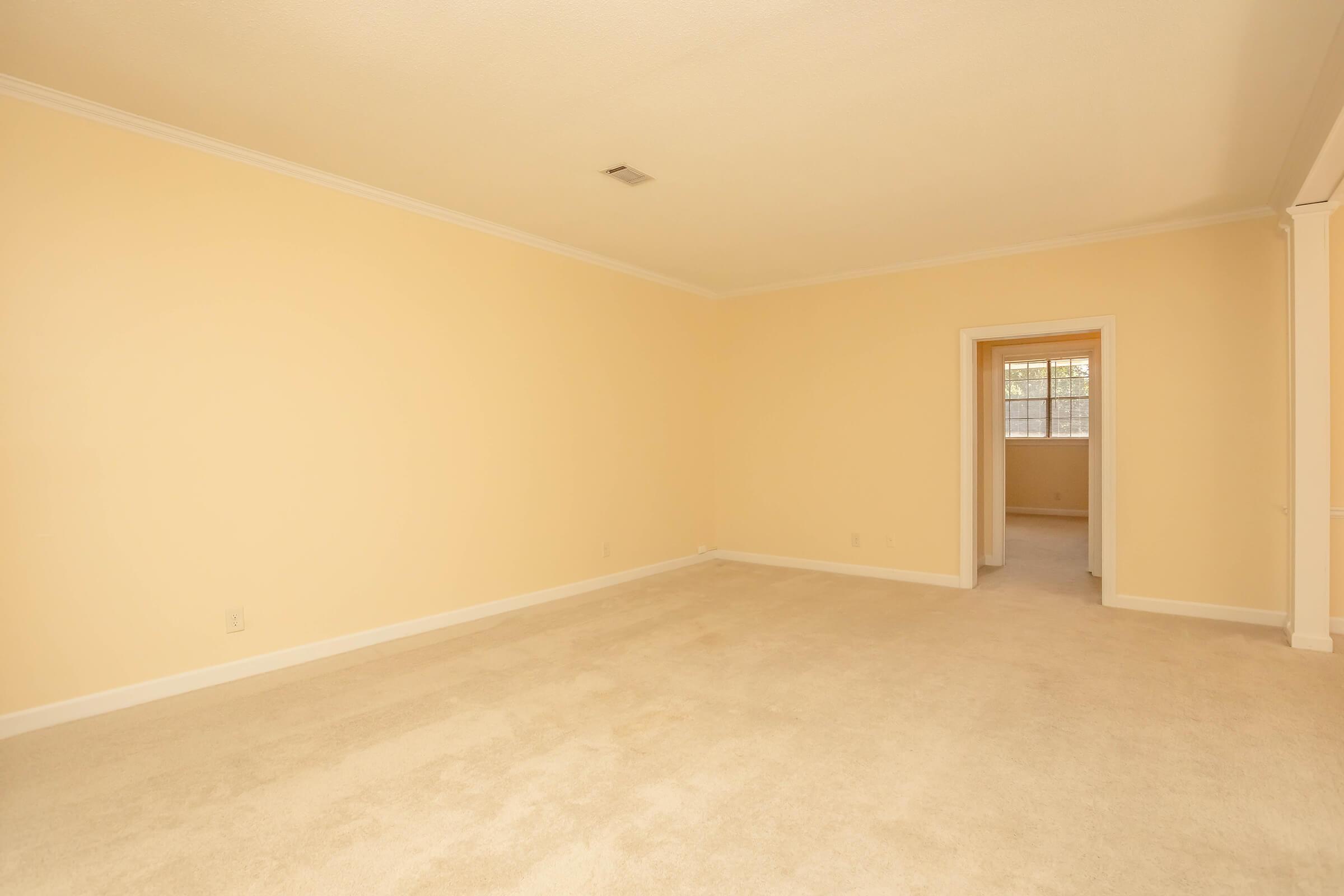 A spacious, empty room with light beige walls and a soft carpeted floor. There is a doorway on the left leading to another room, and a window allowing natural light to enter. The ceiling has a simple crown molding, creating a clean and inviting atmosphere.