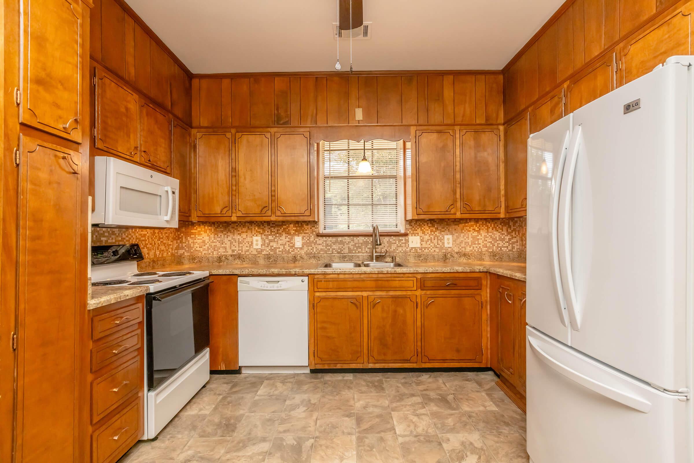 A warm-toned kitchen with wooden cabinets, a granite countertop, and modern appliances including a white refrigerator, dishwasher, oven, and microwave. A window above the sink provides natural light, and the floor has a light-colored tile. The overall ambiance is inviting and cozy.