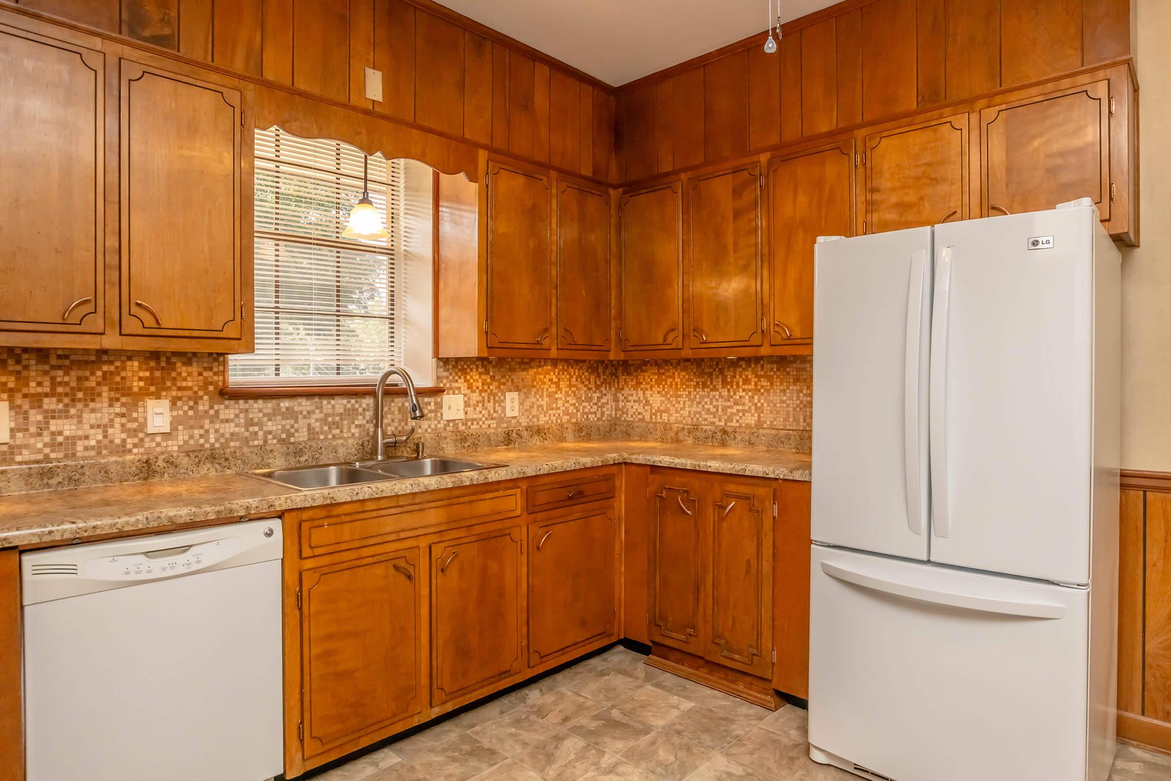 A modern kitchen featuring wooden cabinetry, a double sink with a faucet, a white refrigerator, and a dishwasher. The backsplash consists of small mosaic tiles, and there is natural light from a window above the sink. The flooring is a light-colored vinyl, enhancing the warm tones of the wood.