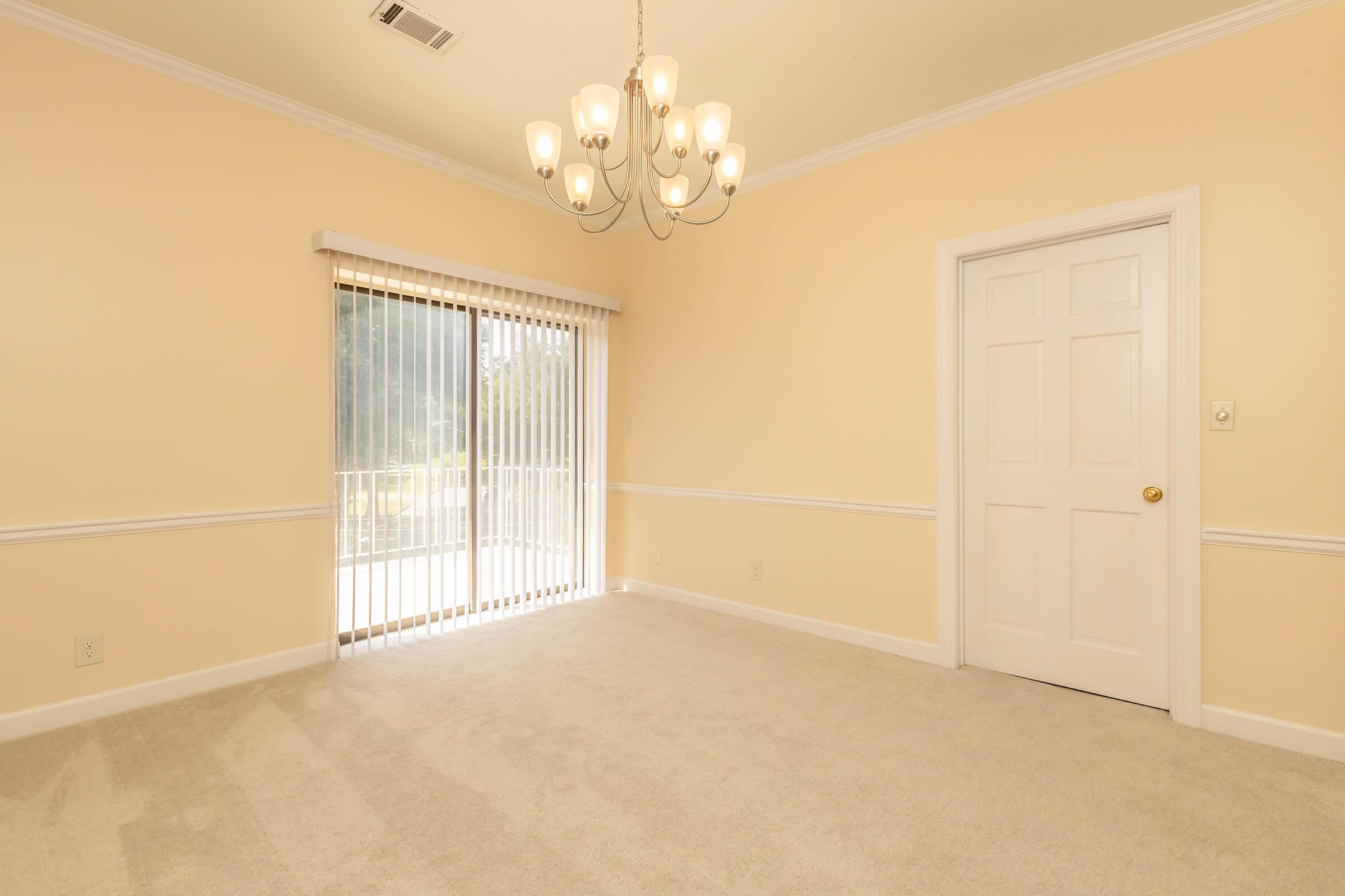A spacious, softly lit room with light beige walls and a plush carpet. A stylish chandelier hangs from the ceiling, and there's a sliding glass door with vertical blinds leading to an outdoor area. A closed white door is visible on the right side of the wall.