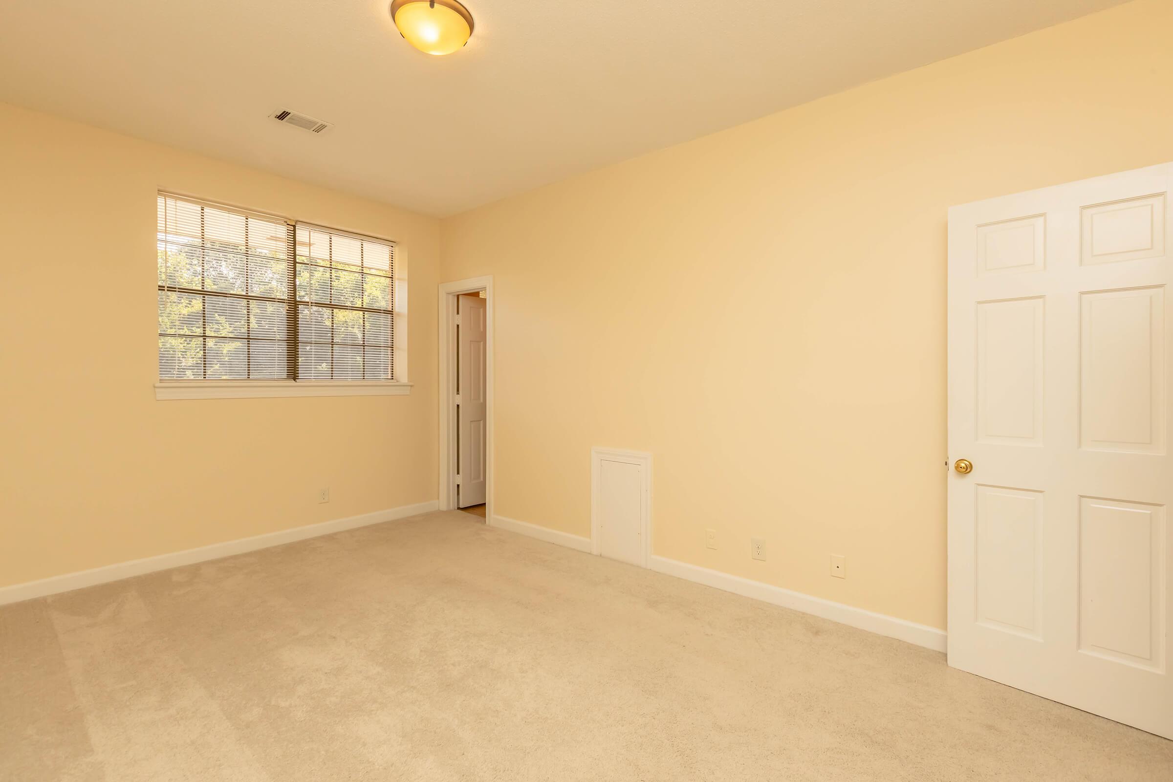 A spacious, empty room with light yellow walls and beige carpet. There is a large window with multiple panes allowing natural light, and a white door on the right leading to another space. The ceiling features a round light fixture. The overall ambiance is bright and clean, suitable for various uses.