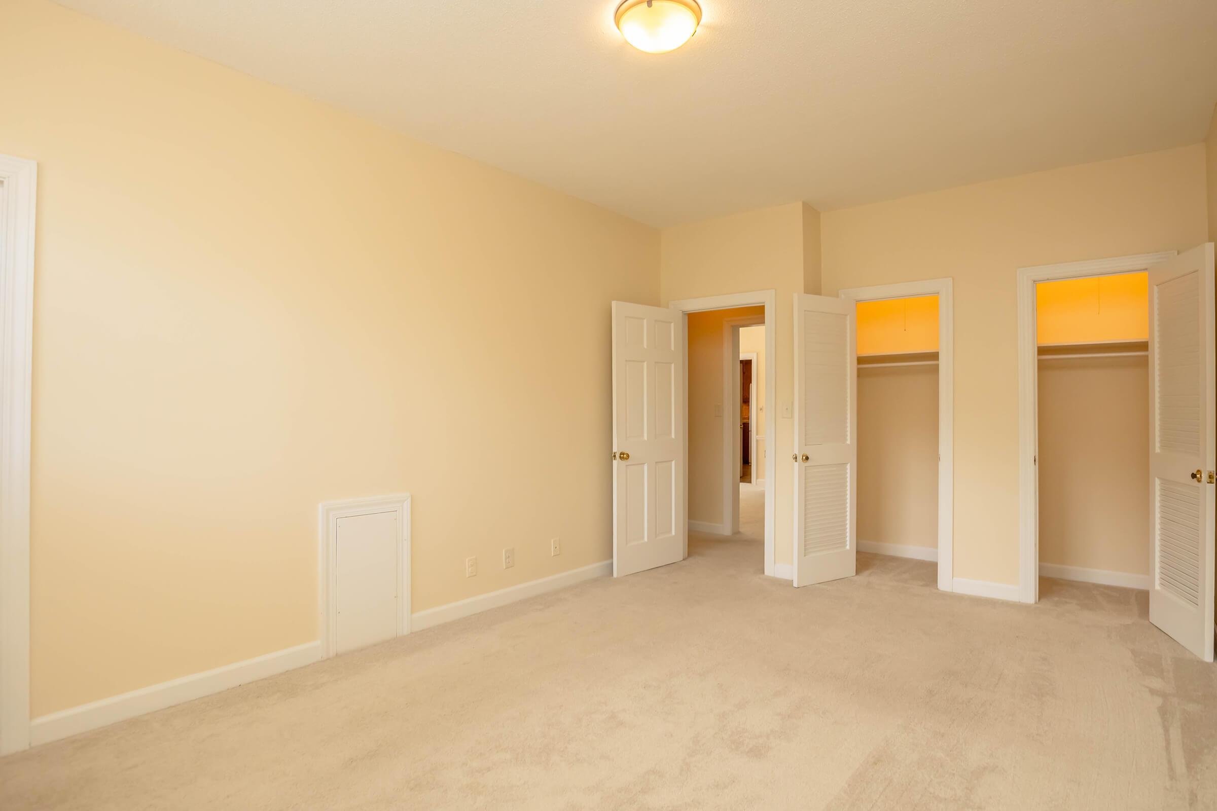 Empty, light-colored bedroom with beige walls and plush carpet. Two open doors lead to adjoining rooms or closets. A ceiling light fixture provides illumination. The space appears clean and spacious, suitable for furnishing and decoration.