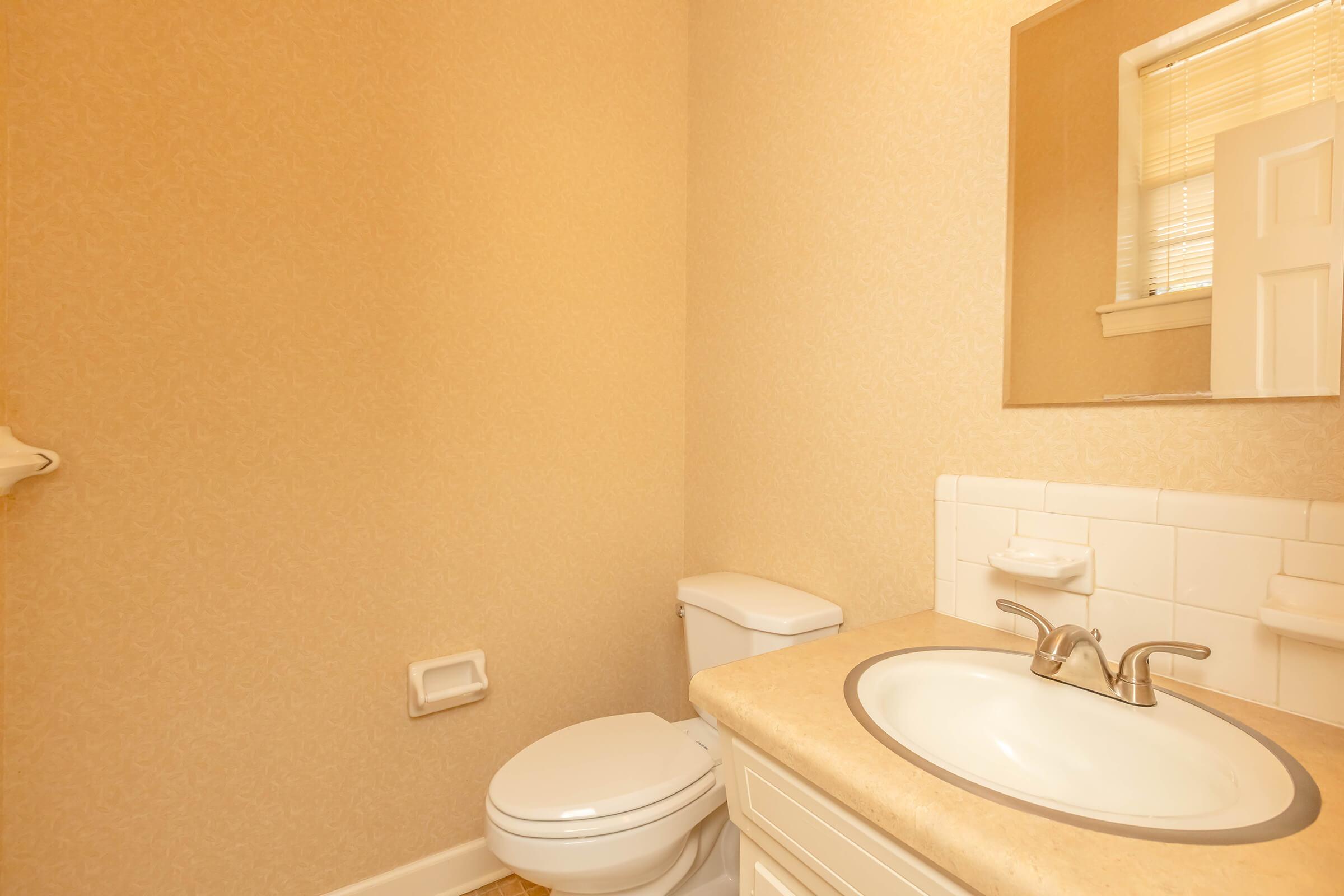 A clean and well-lit bathroom featuring a beige wall, a white toilet, a sink with a round basin, and a wall-mounted mirror above it. The countertop is a light-colored material, and there is a towel bar on the left side. Natural light is coming in from a window.