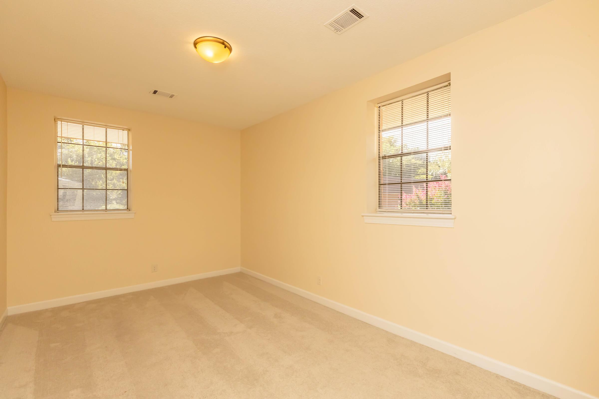 An empty room with light beige walls and two windows, featuring a smooth ceiling with a ceiling light. The floor is covered with light-colored carpet, and there are no furnishings or decorations, creating a bright and spacious feel.