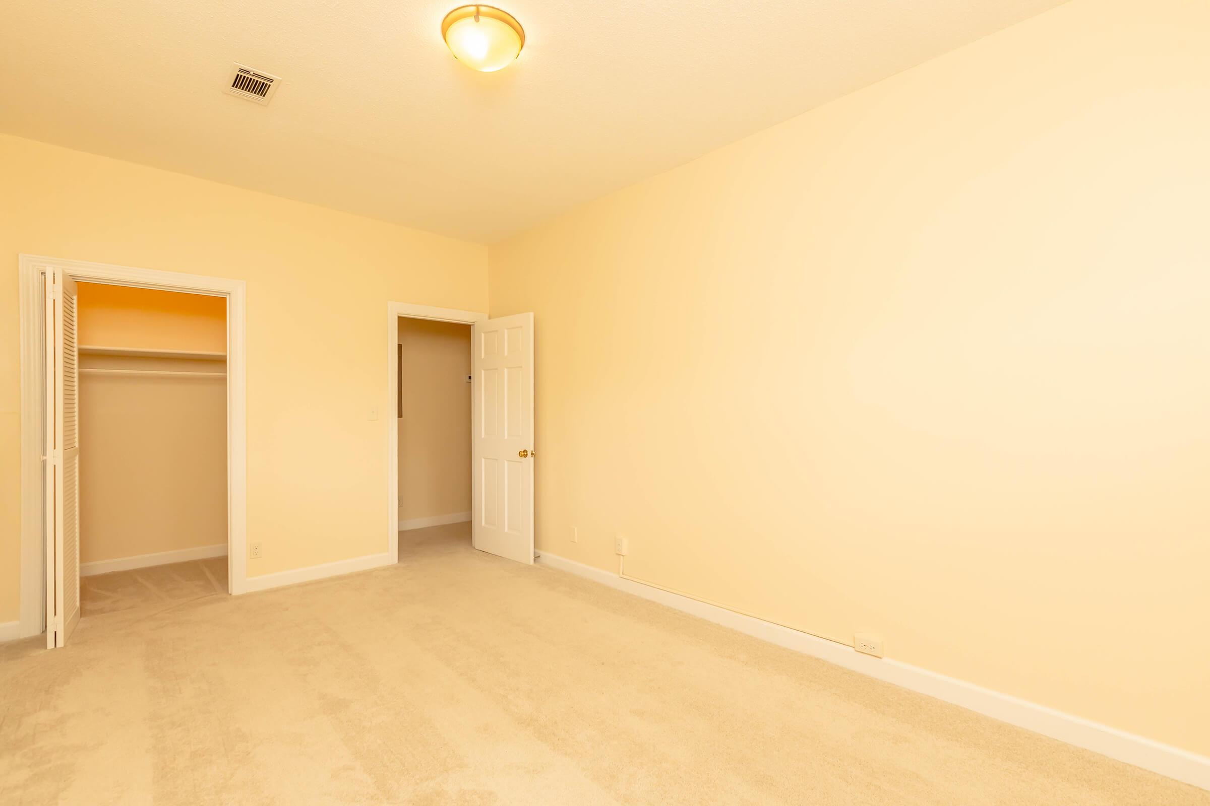 A spacious, empty room with soft beige walls and light carpet. There's a closet on the left with doors open, and a closed door near the back leading to another space. A ceiling light illuminates the area, creating a bright atmosphere. The room appears ready for furnishings.