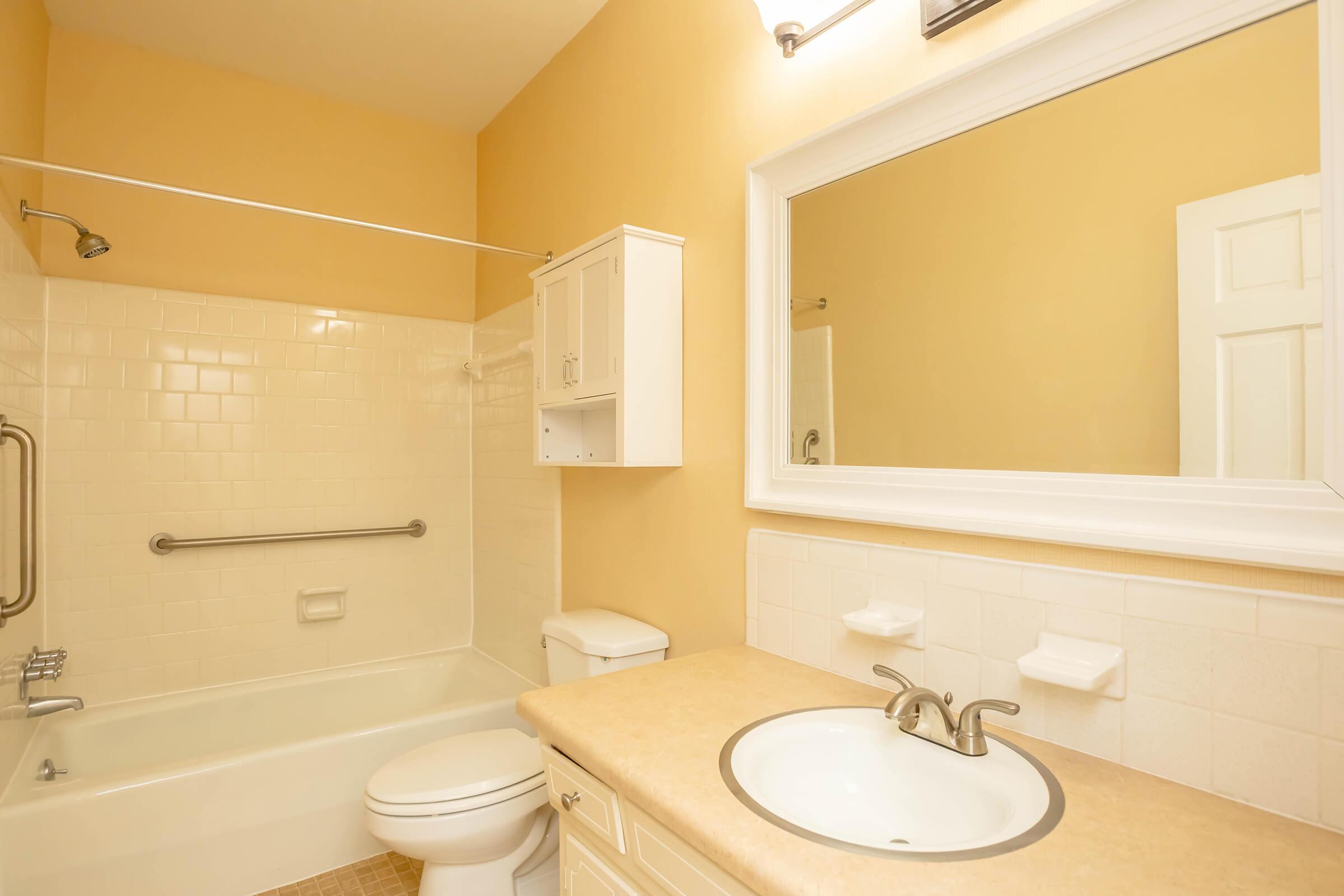 A clean, simple bathroom featuring a bathtub with a shower, a white toilet, a single vanity with a sink, and light-colored walls. A large mirror above the sink adds brightness, while a small cabinet provides storage. The space is well-lit and designed for functionality.