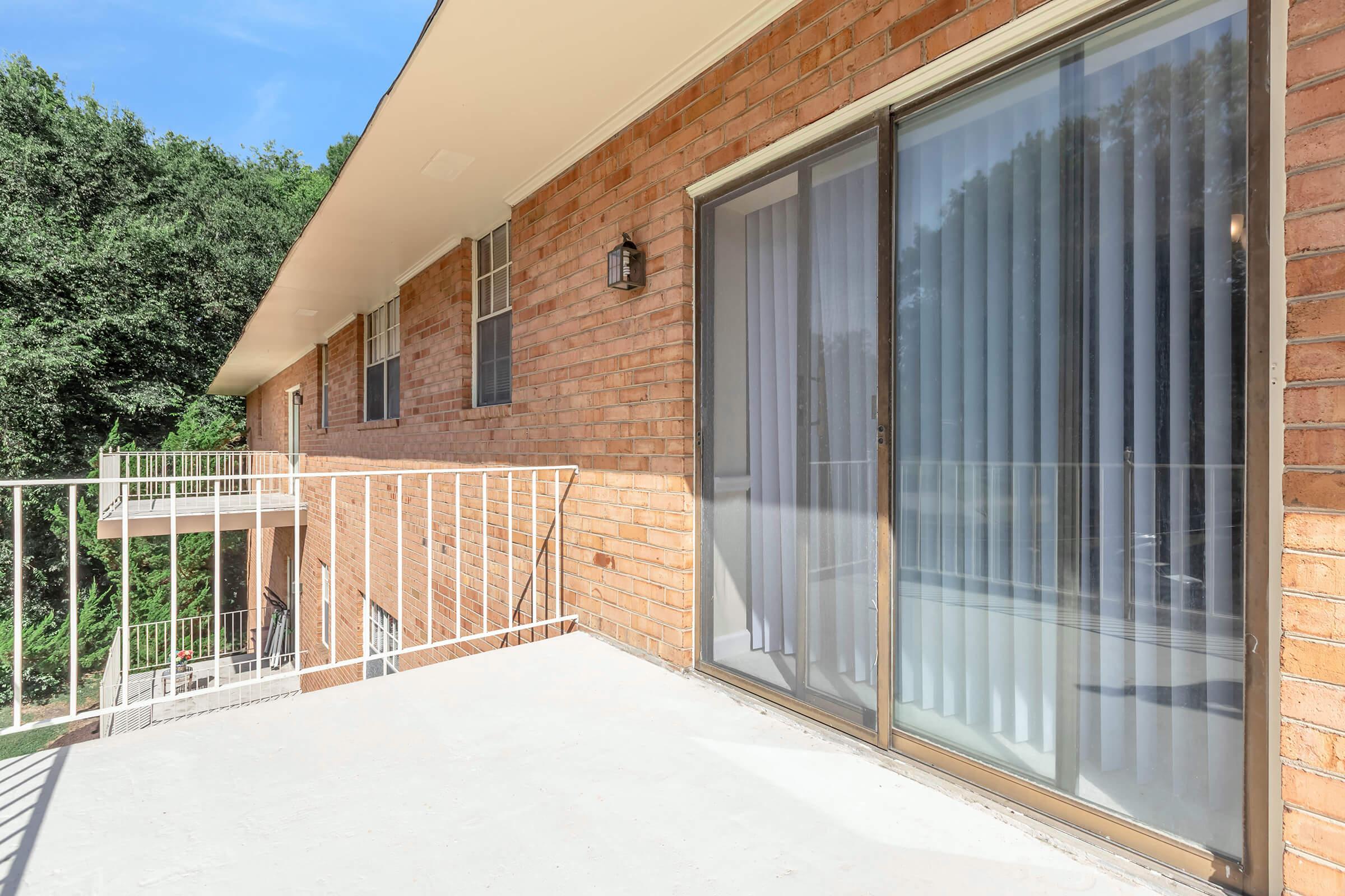 A view of a balcony with a railing, adjacent to a brick building. Large sliding glass doors are visible, leading inside. Lush green trees can be seen in the background under a clear blue sky. The setting is bright and inviting, suggesting a comfortable outdoor space.