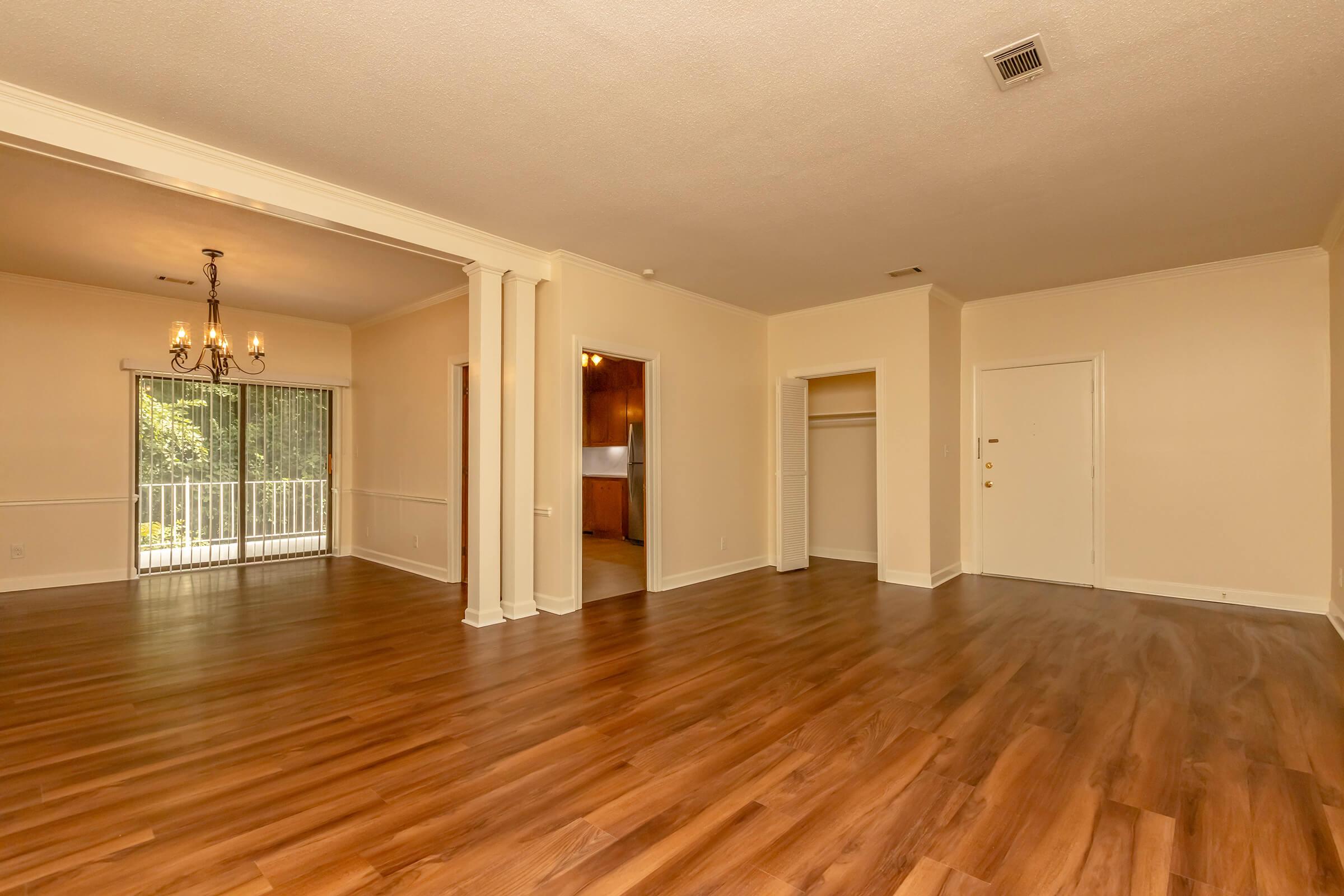Spacious living area with hardwood floor, large windows allowing natural light, and a view of adjacent room featuring a chandelier. A doorway leads to a kitchen area, and a closet is visible to the right. Neutral-colored walls create a warm and inviting atmosphere.