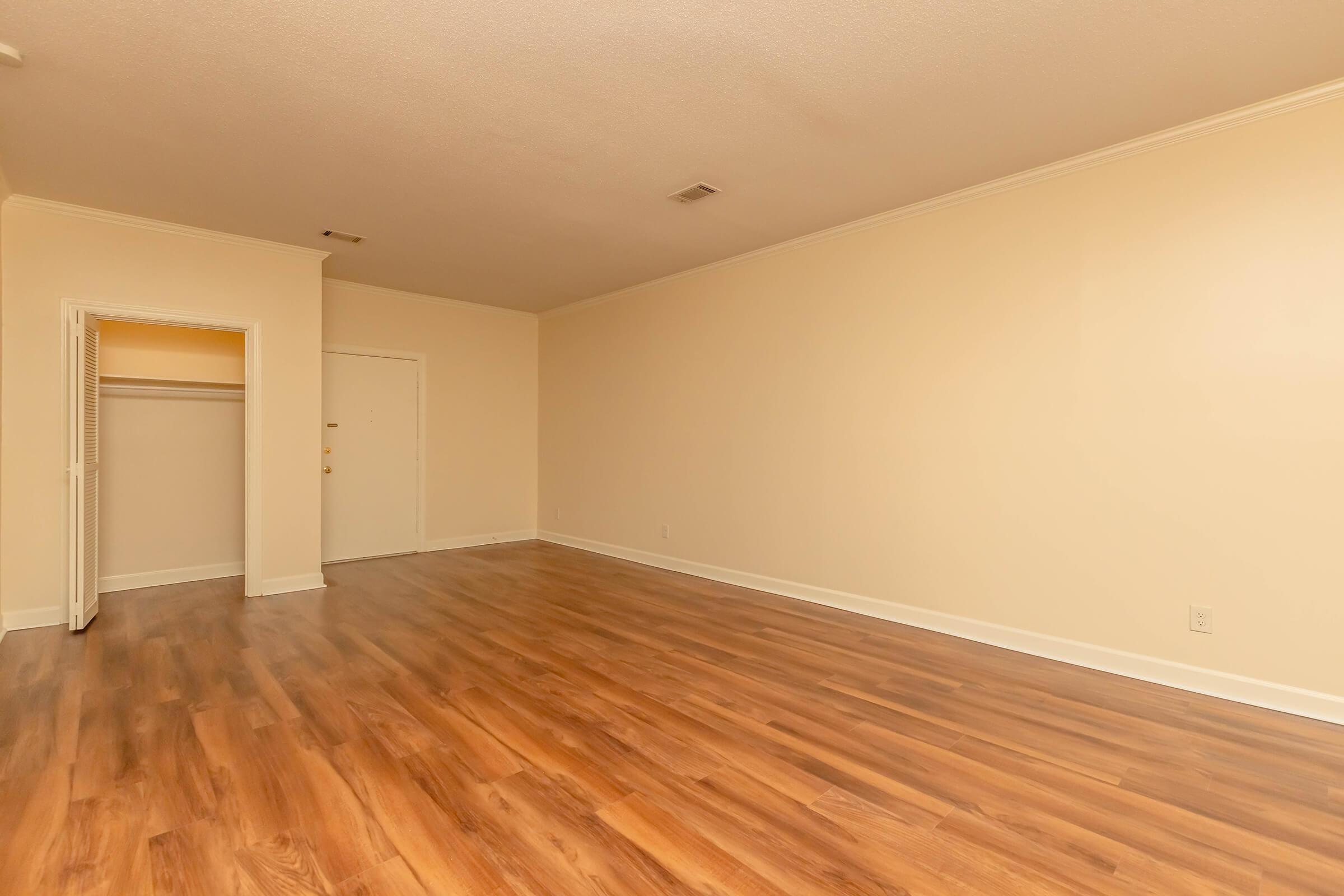 A spacious empty room with light-colored walls and hardwood flooring. There is a closet to the left and a door leading outside. The area is well-lit and ready for furnishing.