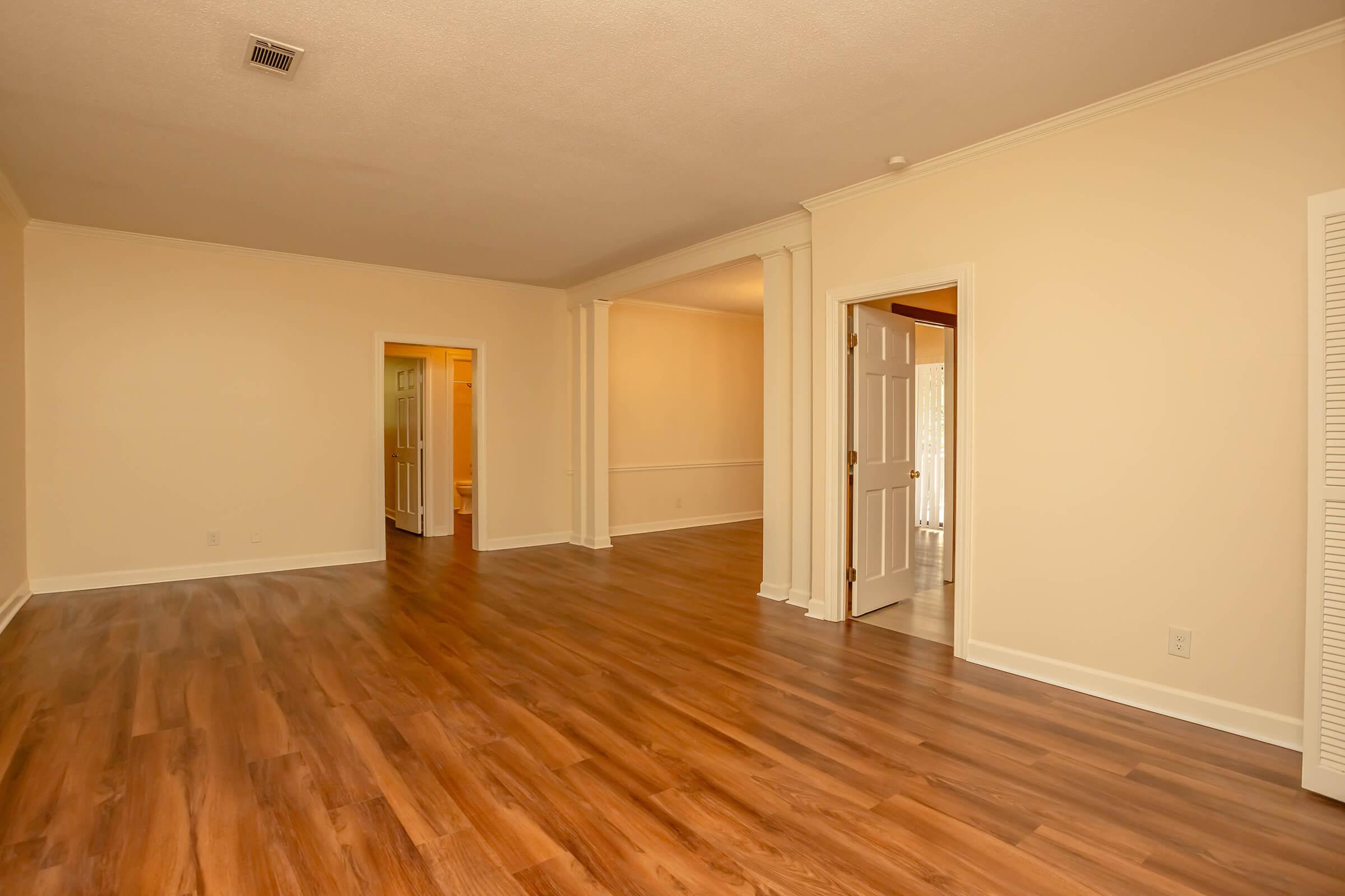A spacious interior room with smooth wooden flooring, light-colored walls, and crown molding. Two doorways visible, one leading to another room and the other to a hallway. Natural light illuminates the space, creating an inviting atmosphere.