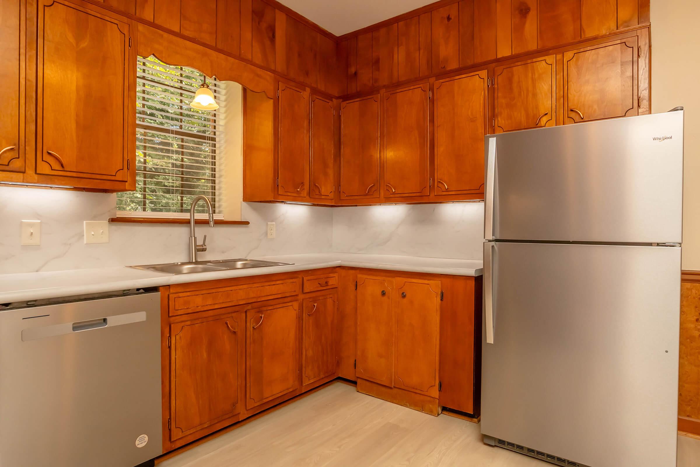 Modern kitchen featuring wooden cabinets, a stainless steel refrigerator, and a sleek silver dishwasher. The countertop is light-colored, and the space is well-lit with natural light coming through a window. It has a neutral color palette and a clean, organized appearance.