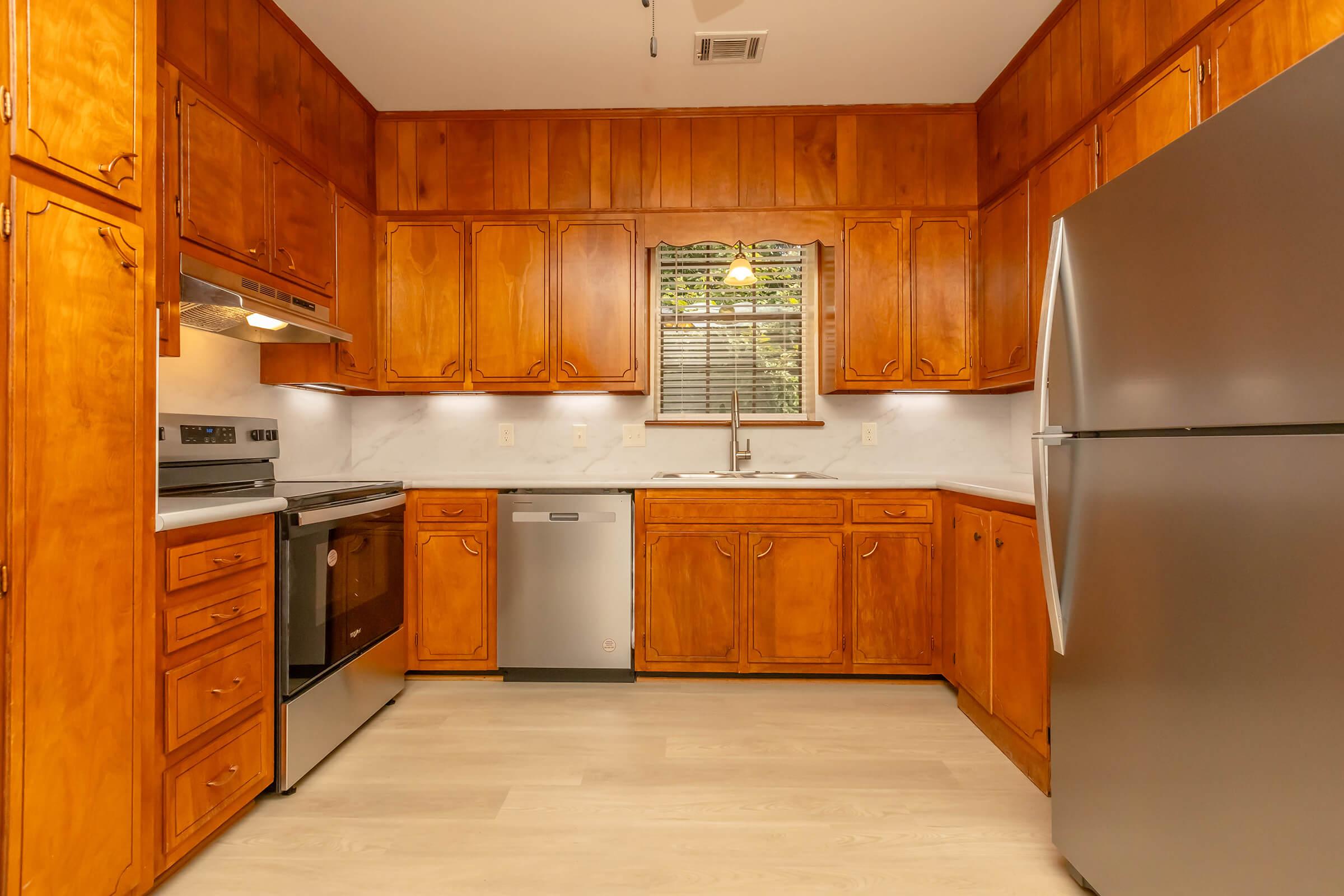 A warm-toned kitchen featuring wooden cabinets, a stainless steel refrigerator, a modern stove, a built-in dishwasher, and a sink with a window above it. The countertop is light-colored, and the flooring is a pale shade, creating a cozy and inviting atmosphere.