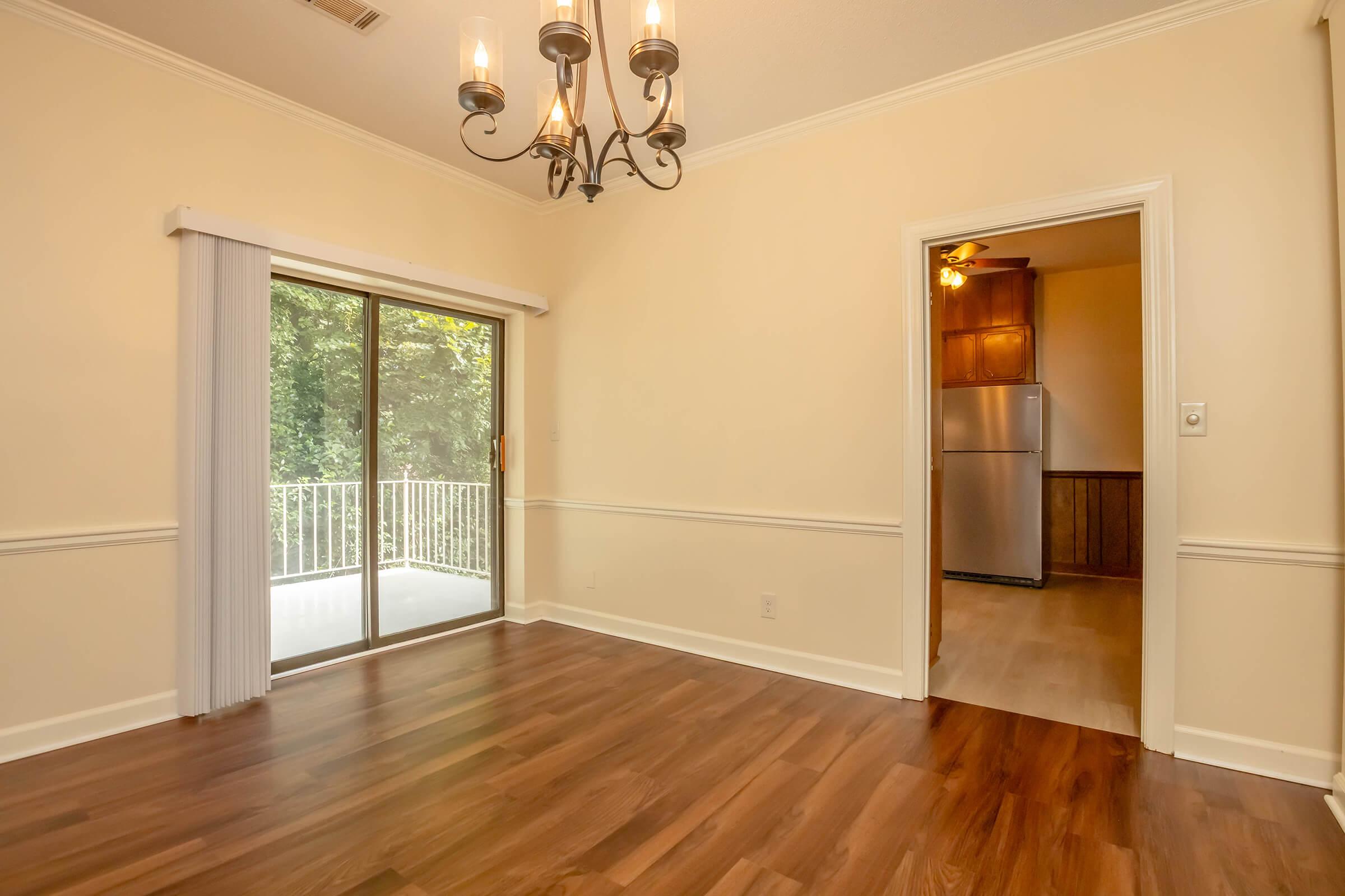 A well-lit room with wooden flooring, featuring a ceiling chandelier. There is a sliding glass door leading to a balcony, and an adjoining doorway that leads to a kitchen area. The walls are painted a light color, and there are decorative moldings along the bottom.