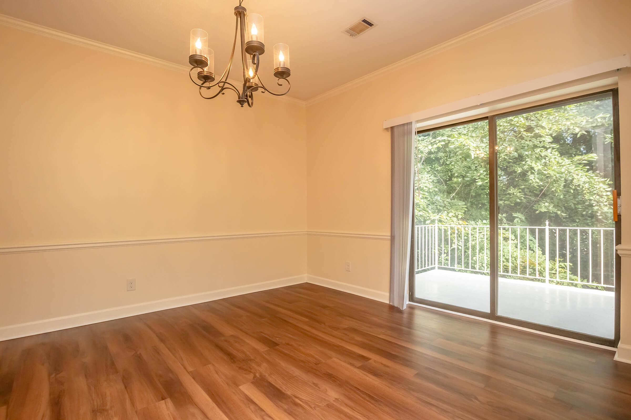 A bright, empty room featuring hardwood flooring, a chandelier, and a large sliding glass door that opens to a balcony with views of trees outside. The walls are painted a soft yellow, and there is a decorative white molding along the lower part of the walls.