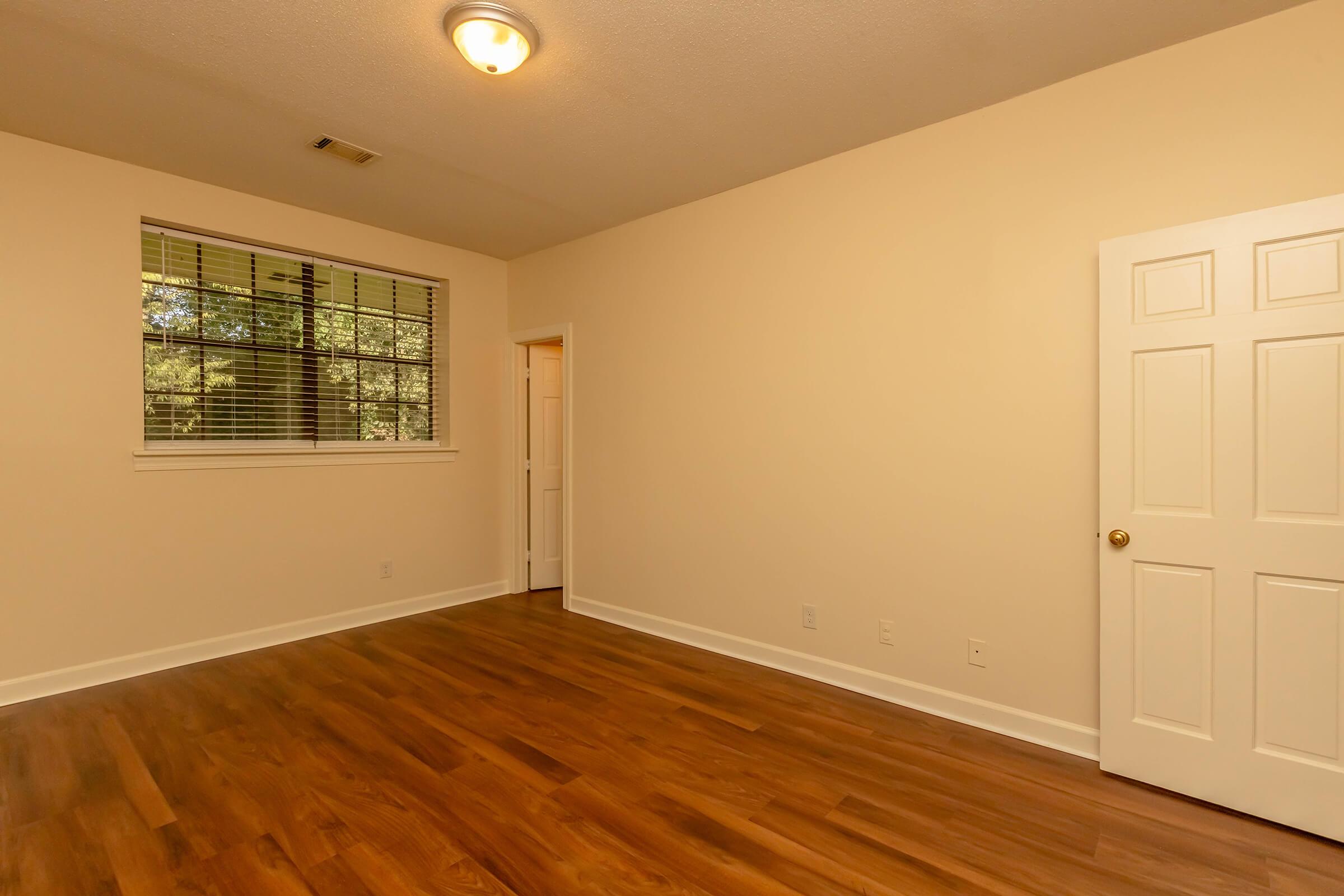 A spacious, empty room with light-colored walls and hardwood flooring. It features a window with blinds, providing natural light. A closed door is on the right, and the room has a ceiling light fixture. The overall atmosphere is bright and inviting, suitable for personal decoration or use.