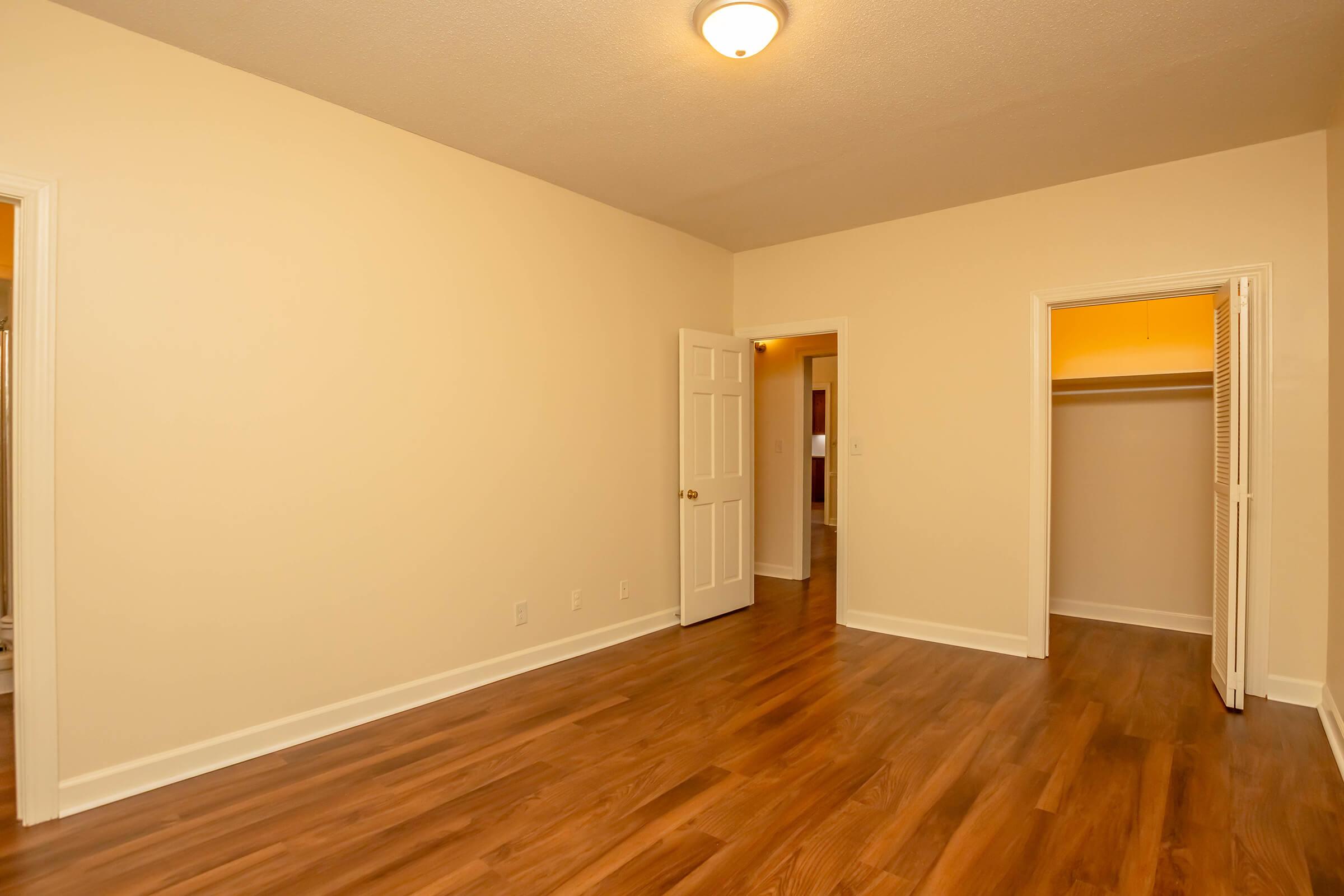 Empty room with light beige walls and a hardwood floor. One door leads to a closet, and another door opens to a hallway. The ceiling light provides illumination, highlighting the spaciousness of the area. The overall appearance is clean and freshly painted.