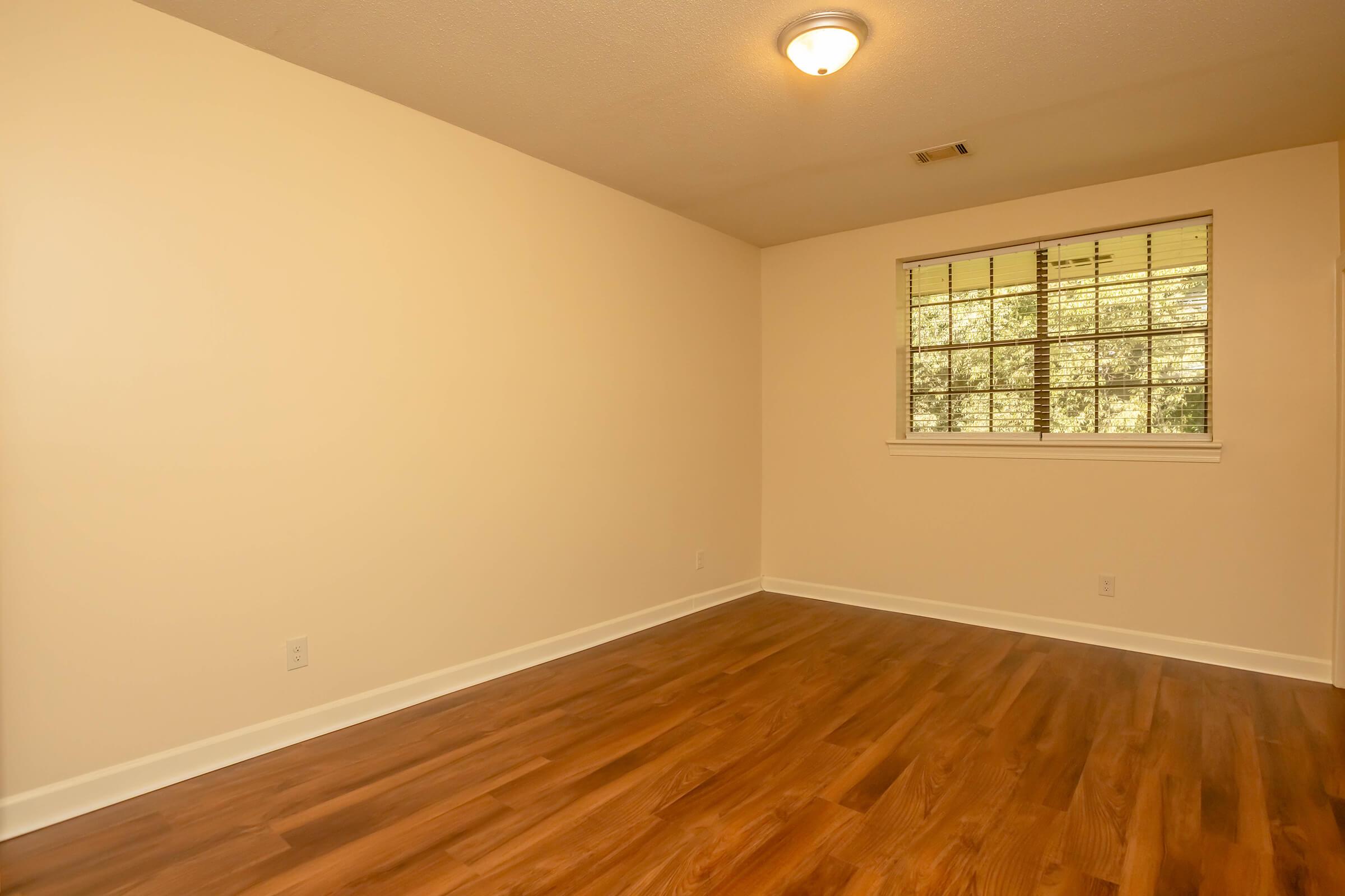 Empty room with light-colored walls and a wooden floor. A window with blinds lets in natural light, and a ceiling light fixture is visible. The space appears tidy and unfurnished, suitable for various uses such as a living area or bedroom.