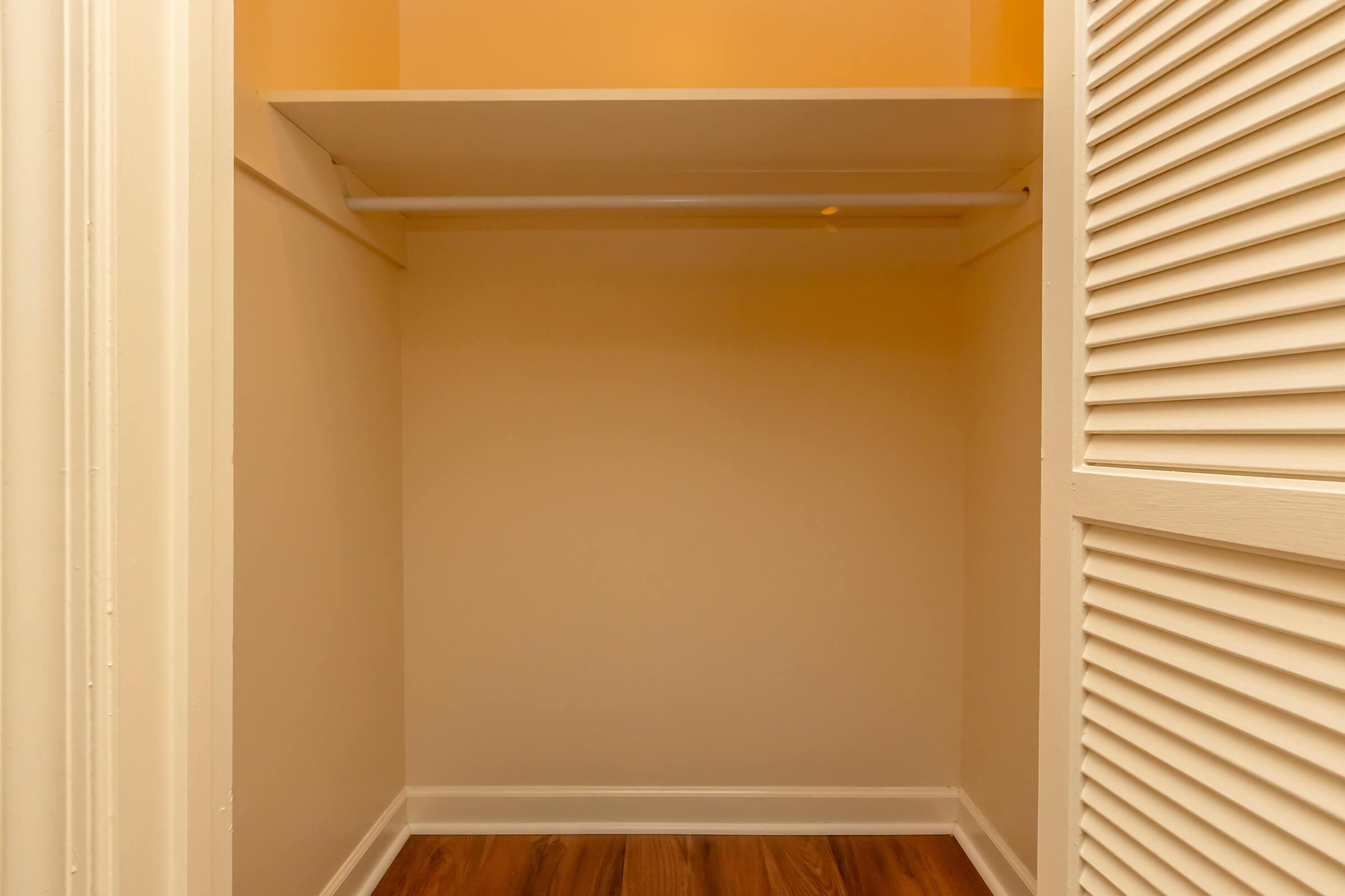 Empty closet space with a single shelf and a hanging rod, featuring light-colored walls and a wooden floor. The closet door is open, revealing the simplicity of the interior design.