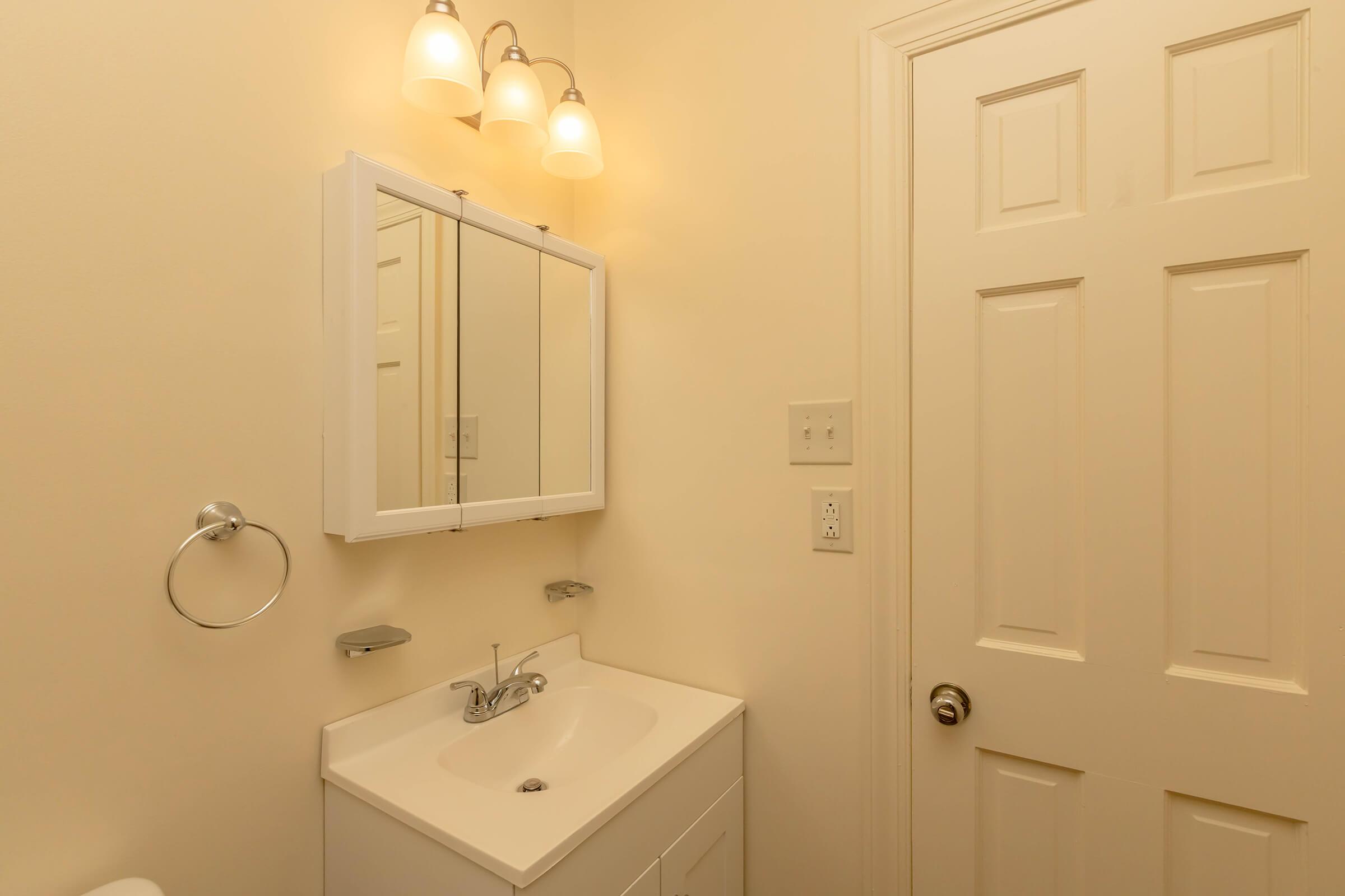 A bright bathroom featuring a white vanity with a sink, a mirror cabinet above, and soft lighting from three fixtures. The walls are a light cream color, and a closed door is visible on the right, adding to the overall clean and minimalist look.