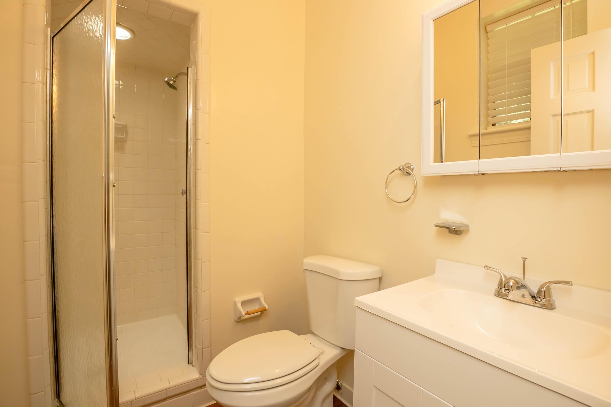 A bright, clean bathroom featuring a small shower with a glass door, a modern toilet, and a white vanity with a sink. The walls are painted a light yellow, and there is a mirror above the sink. Natural light enters through a window with blinds. The overall aesthetic is simple and inviting.