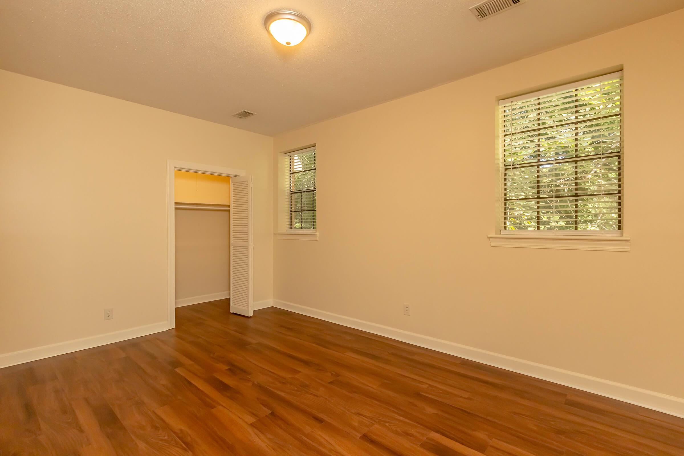 Empty room with light wooden floors, beige walls, and two windows with white blinds. There is a closet with sliding doors on one side, and overhead light fixture. Natural light enters the room, creating a bright and airy atmosphere.