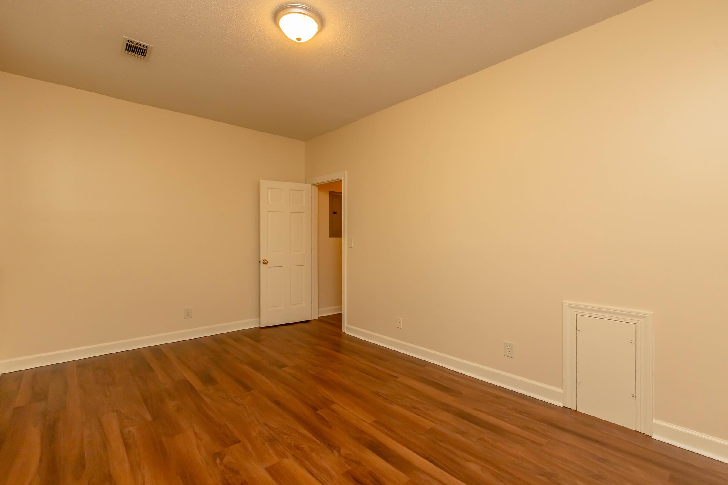 Empty room with light-colored walls and a hardwood floor. A single light fixture is visible on the ceiling, and there are two doors: one leading to another room and the other with a small opening near the floor, possibly a vent or storage access. The space is well-lit and uncluttered.
