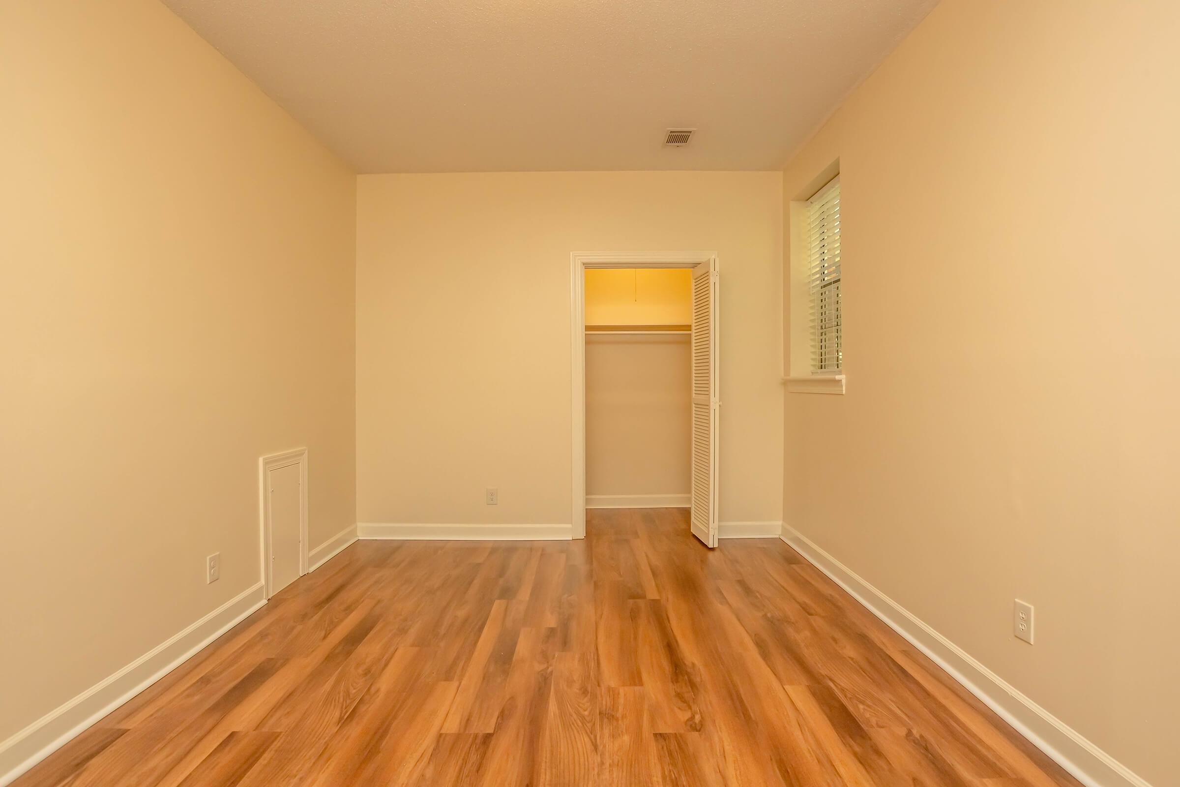 A vacant room with light beige walls, wood-like flooring, and a small closet. Natural light enters through a window. The room has a simple and empty appearance, suitable for potential furnishings and personal decoration.