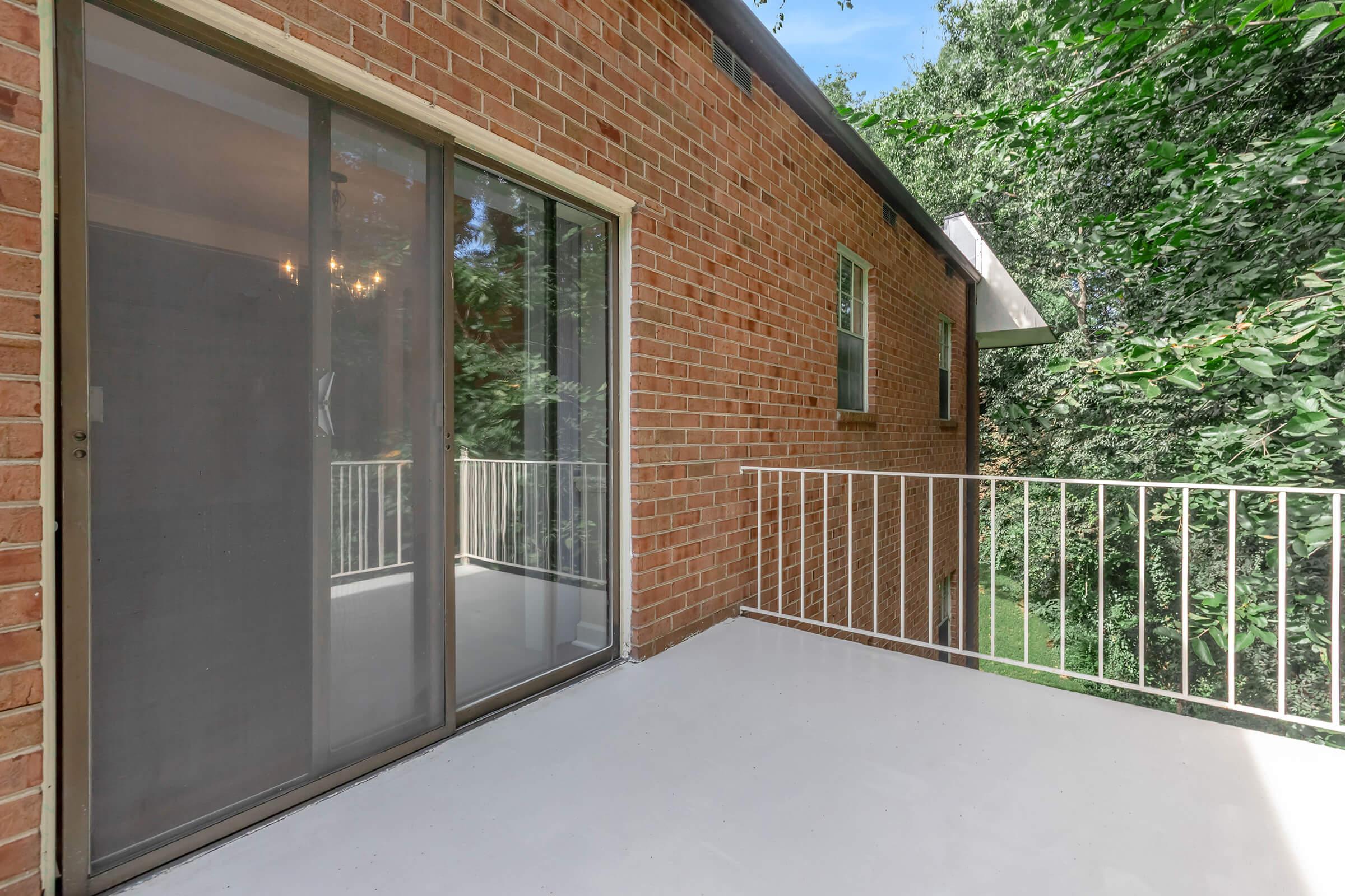 A small balcony with a white railing, featuring a sliding glass door. The exterior wall is made of brick, and trees are visible in the background, creating a natural setting. The balcony floor is smooth and painted white, suggesting a clean and open space.
