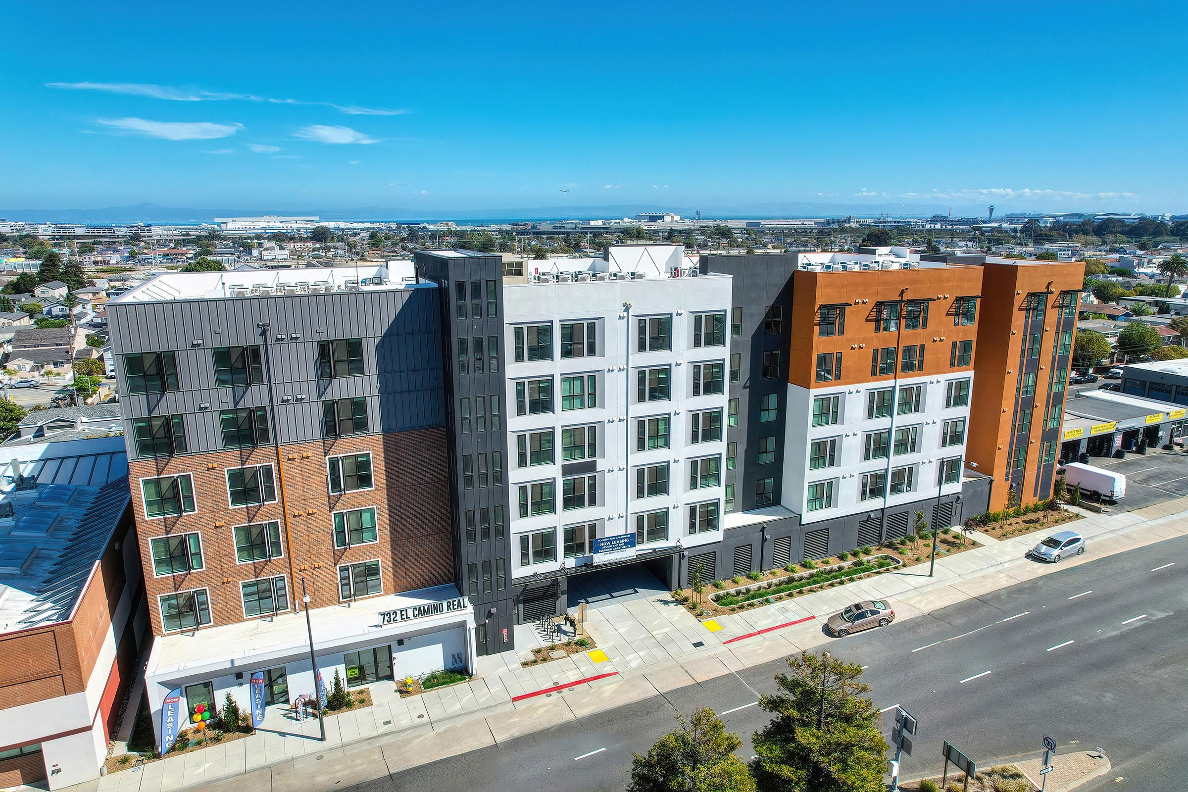 Aerial view of a modern multi-story apartment building featuring a mix of brick and contemporary architecture. The scene includes a clear blue sky, parked cars along the street, and landscaped surroundings, with additional buildings visible in the background.