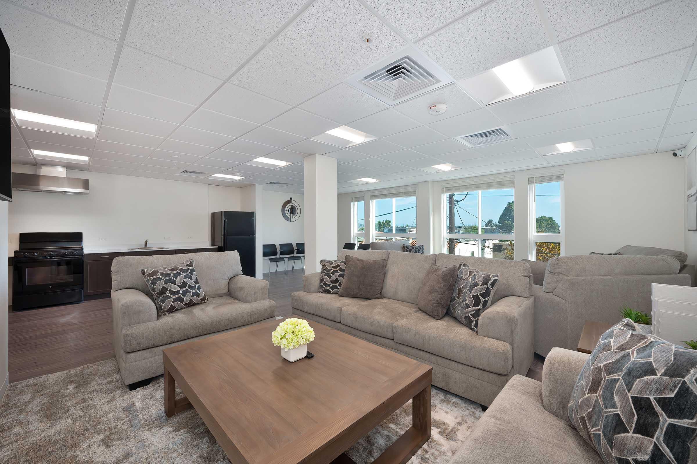 Modern living room with light gray sofas and decorative pillows arranged around a wooden coffee table. A neutral-colored rug is underfoot. In the background, there is a kitchen area with dark cabinets and black appliances, as well as large windows allowing natural light to enter the space.
