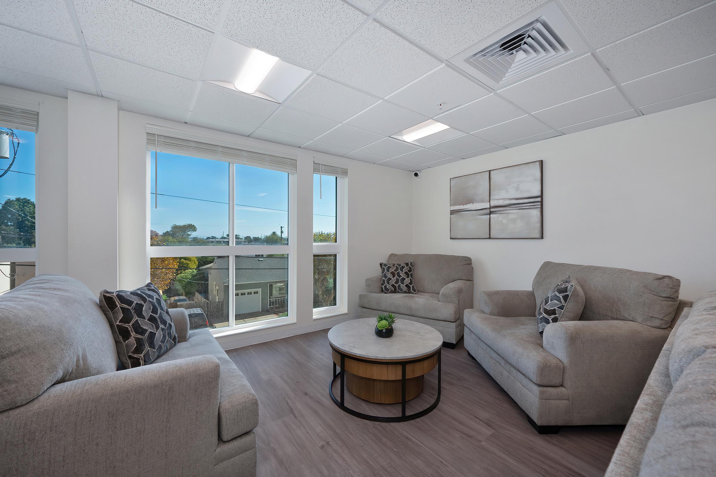 A bright and modern sitting area featuring two light gray sofas with decorative black and white pillows, arranged around a round wooden table. Large windows allow natural light to fill the space, offering a view of greenery outside. The walls are painted white with a simple abstract art piece.