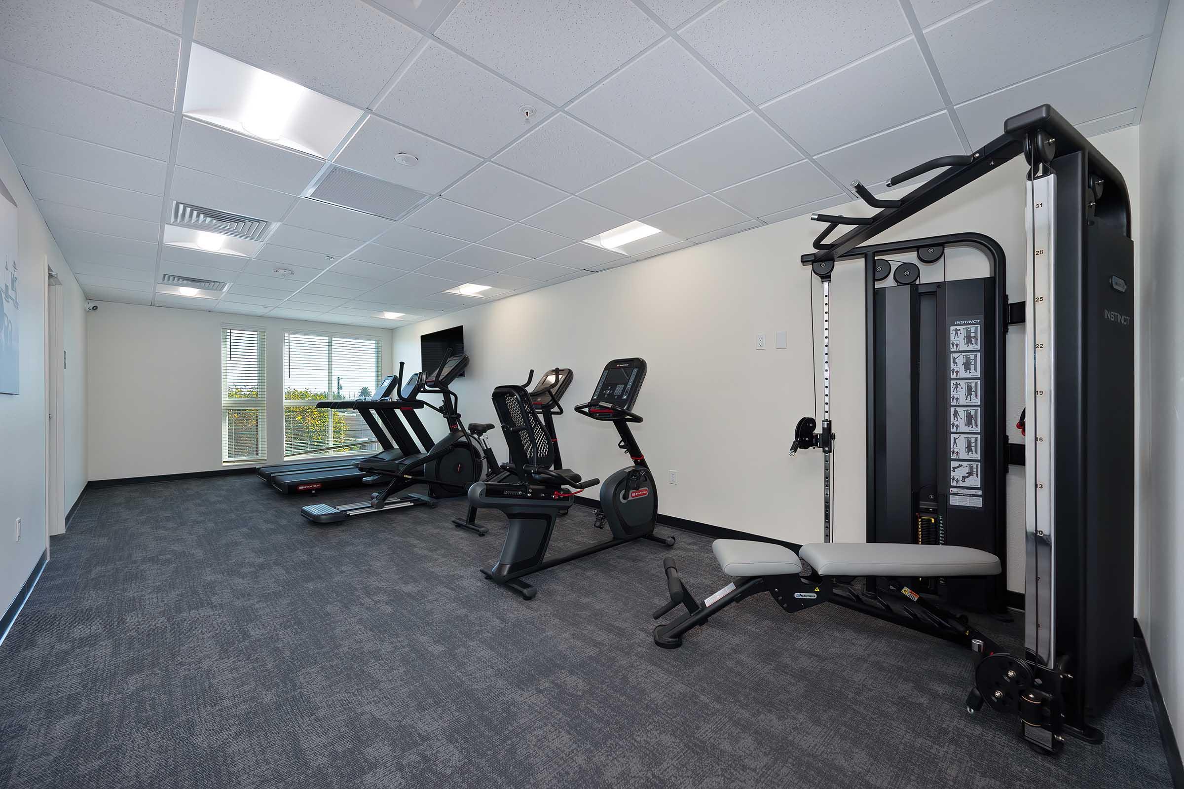 A modern fitness room featuring various exercise equipment, including treadmills, elliptical machines, and a multi-station weight machine. The space has bright lighting and large windows allowing natural light, enhancing the workout environment.