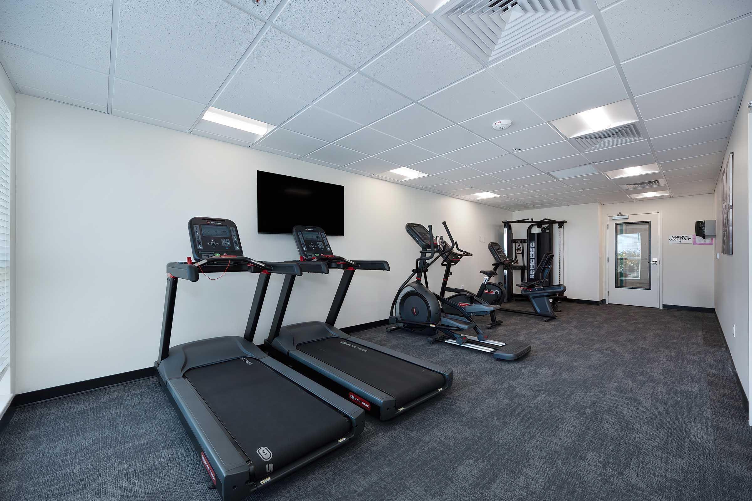 A modern fitness room featuring two treadmills, an elliptical machine, a multi-station weight machine, and a wall-mounted TV. The space has a neutral color scheme with gray carpeting and bright overhead lighting, creating a clean and inviting workout environment.