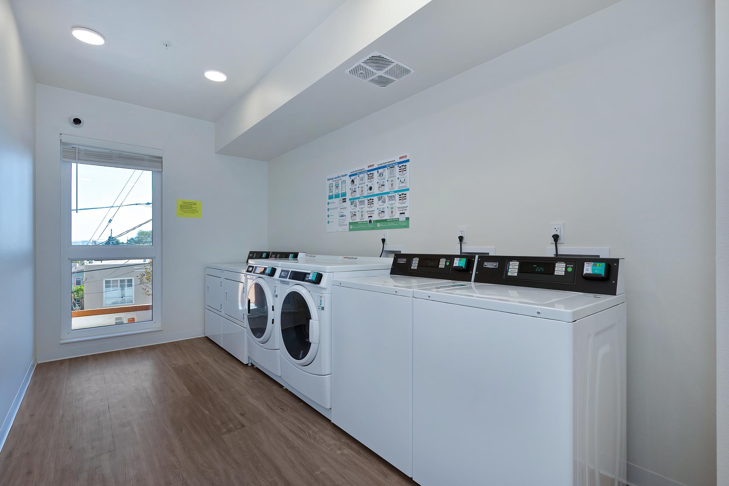 A clean laundry room featuring multiple white washers and dryers lined up against a wall. A window allows natural light to enter, with a view outside. On the wall, a poster provides laundry instructions and information. The room has a modern design with light wood flooring.