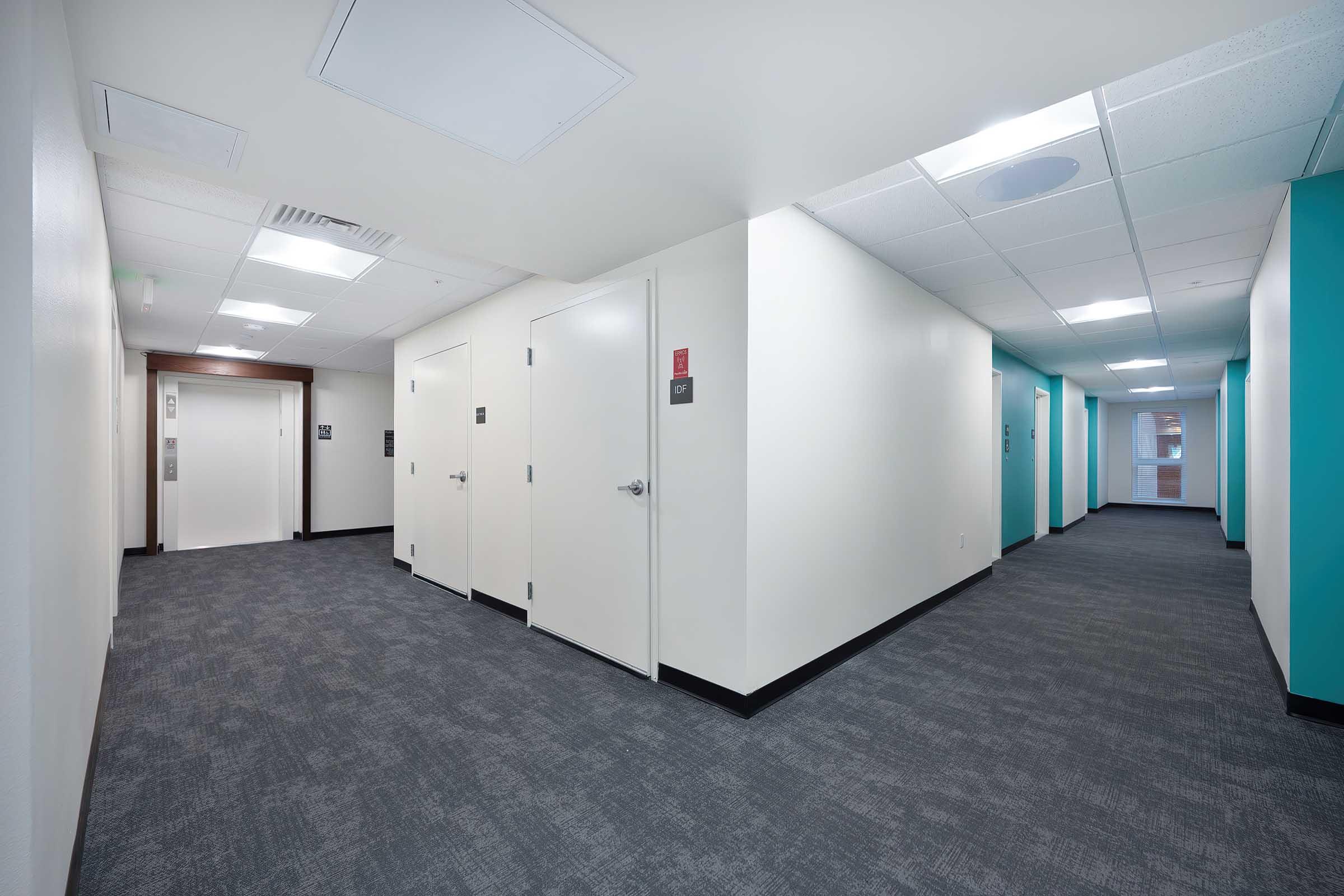 A modern hallway with light-colored walls and dark carpet. The corridor features several closed doors on each side, interspersed with elevator access points. Bright overhead lighting illuminates the space, and there are teal accents on the walls at the end of the hallway.