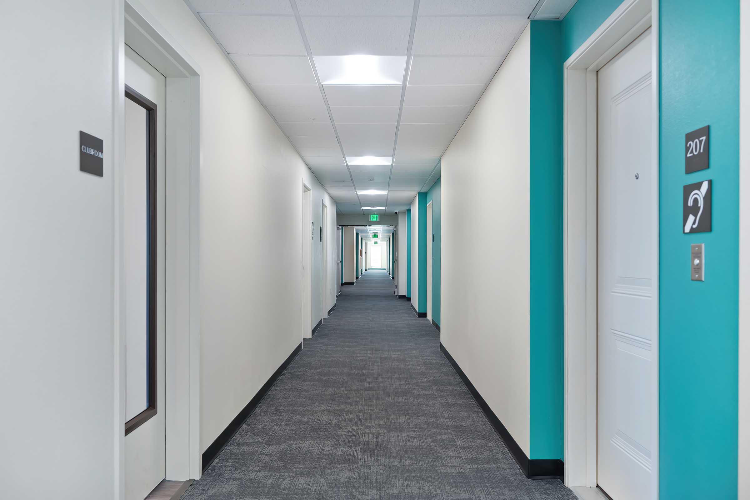 A clean, modern hotel corridor featuring beige walls and turquoise accents. Multiple doors line the hallway, with room numbers visible. The floor is carpeted in dark gray, and bright fluorescent lights illuminate the space, creating a welcoming atmosphere.