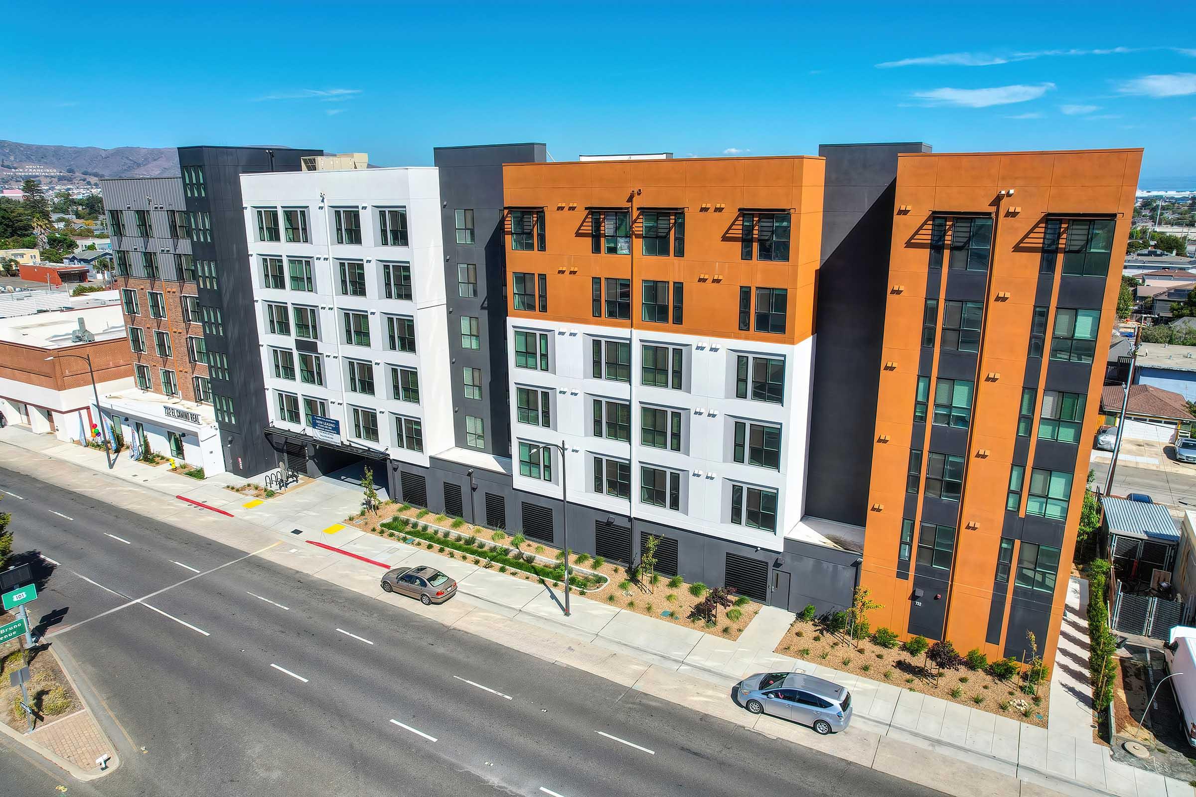 A modern multi-story residential building featuring a mix of white, orange, and gray exterior panels, situated along a city street. The structure has large windows, landscaping in front, and is surrounded by urban surroundings with a clear blue sky above.