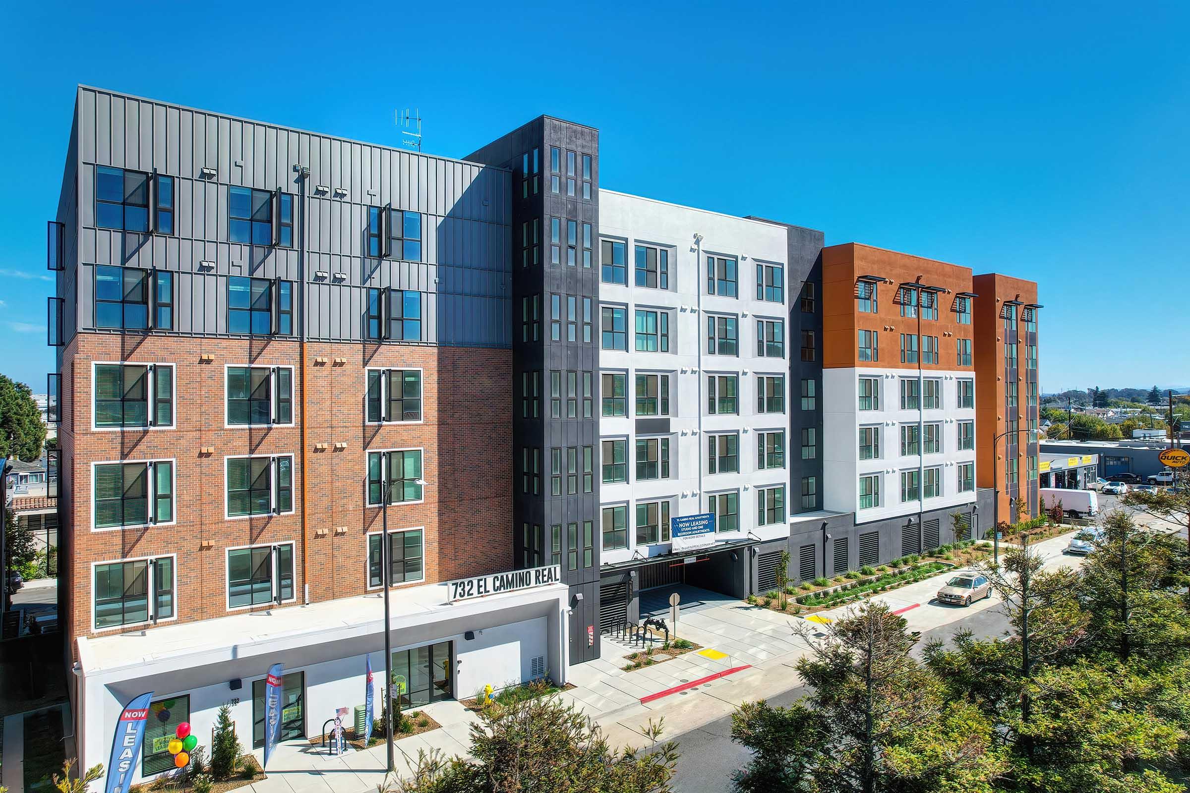Modern multi-story apartment building featuring a mix of brick and contemporary paneling. The structure has large windows and balconies, surrounded by landscaping and parking spaces. Clear blue sky in the background enhances the urban setting.