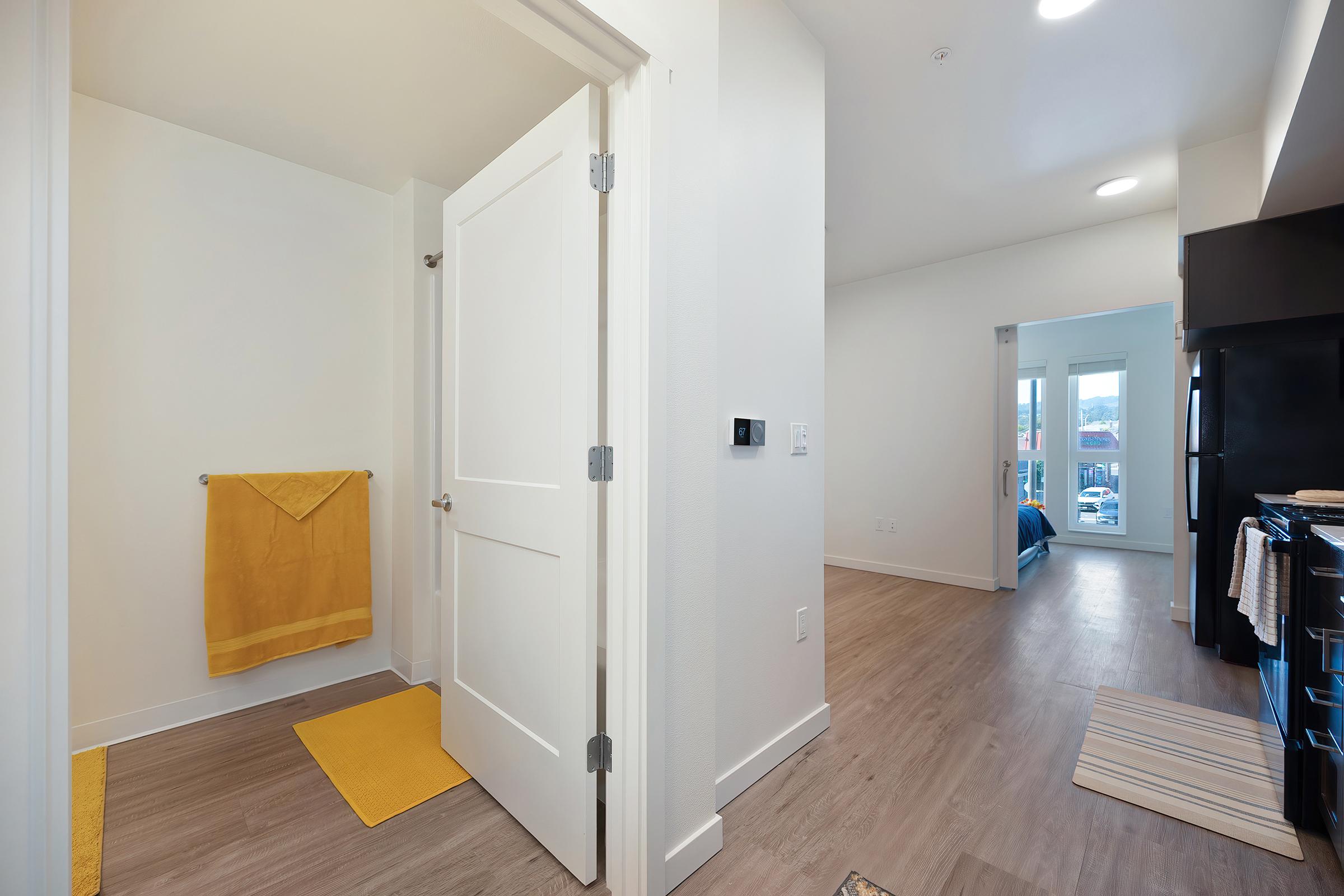 A bright and modern apartment hallway leads to a doorway with a white door on the left, featuring a yellow towel and mat. The room is well-lit with natural light shining through an adjacent room with a blue sofa visible. The flooring is light wood, adding to the contemporary feel of the space.