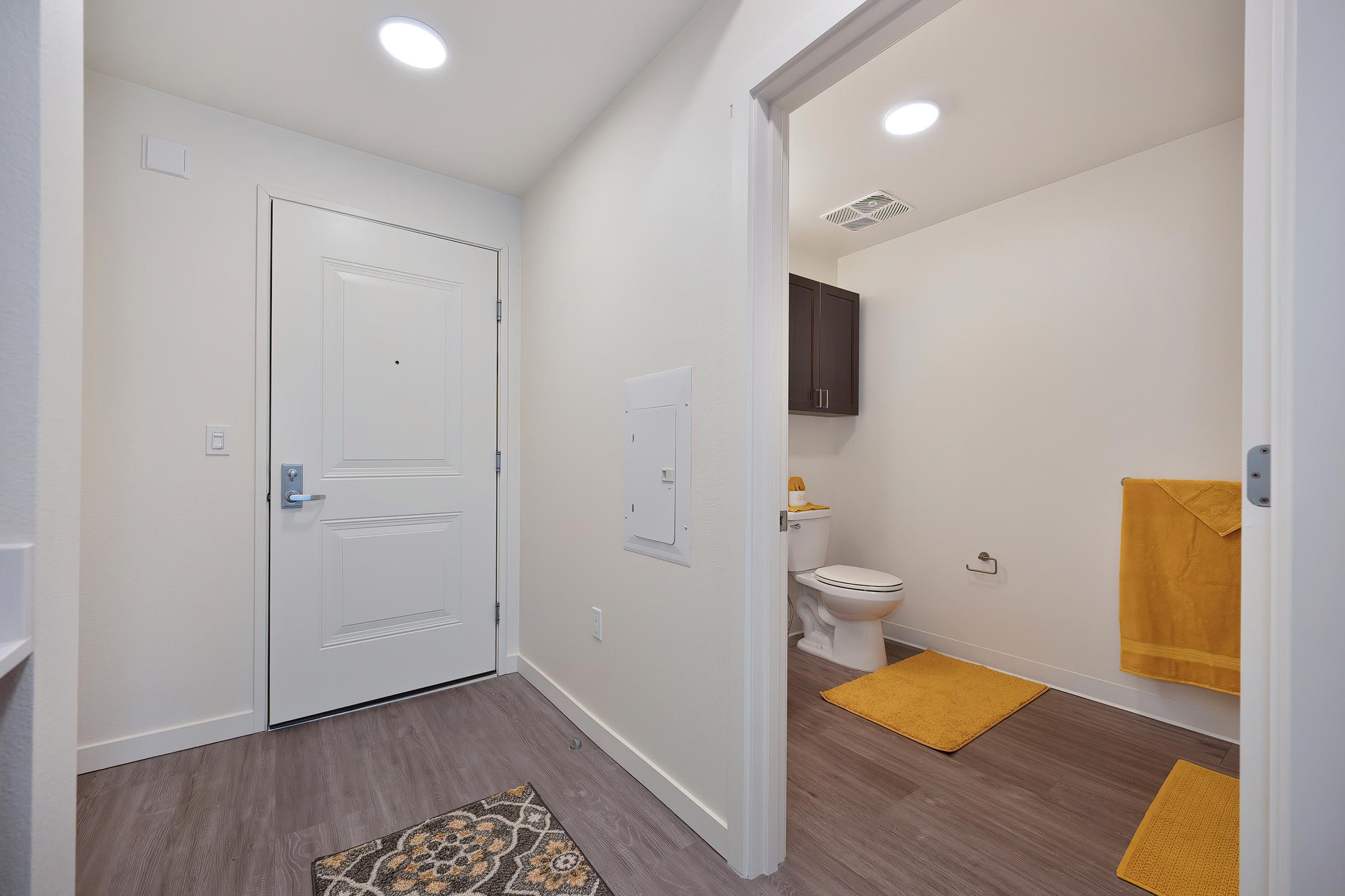 A well-lit entryway leads to a bathroom. The bathroom features a toilet and yellow bath towels, with matching bath mats on the floor. The walls are painted white, and there is a decorative rug at the entrance. Modern lighting fixtures are installed on the ceiling, enhancing the inviting atmosphere.
